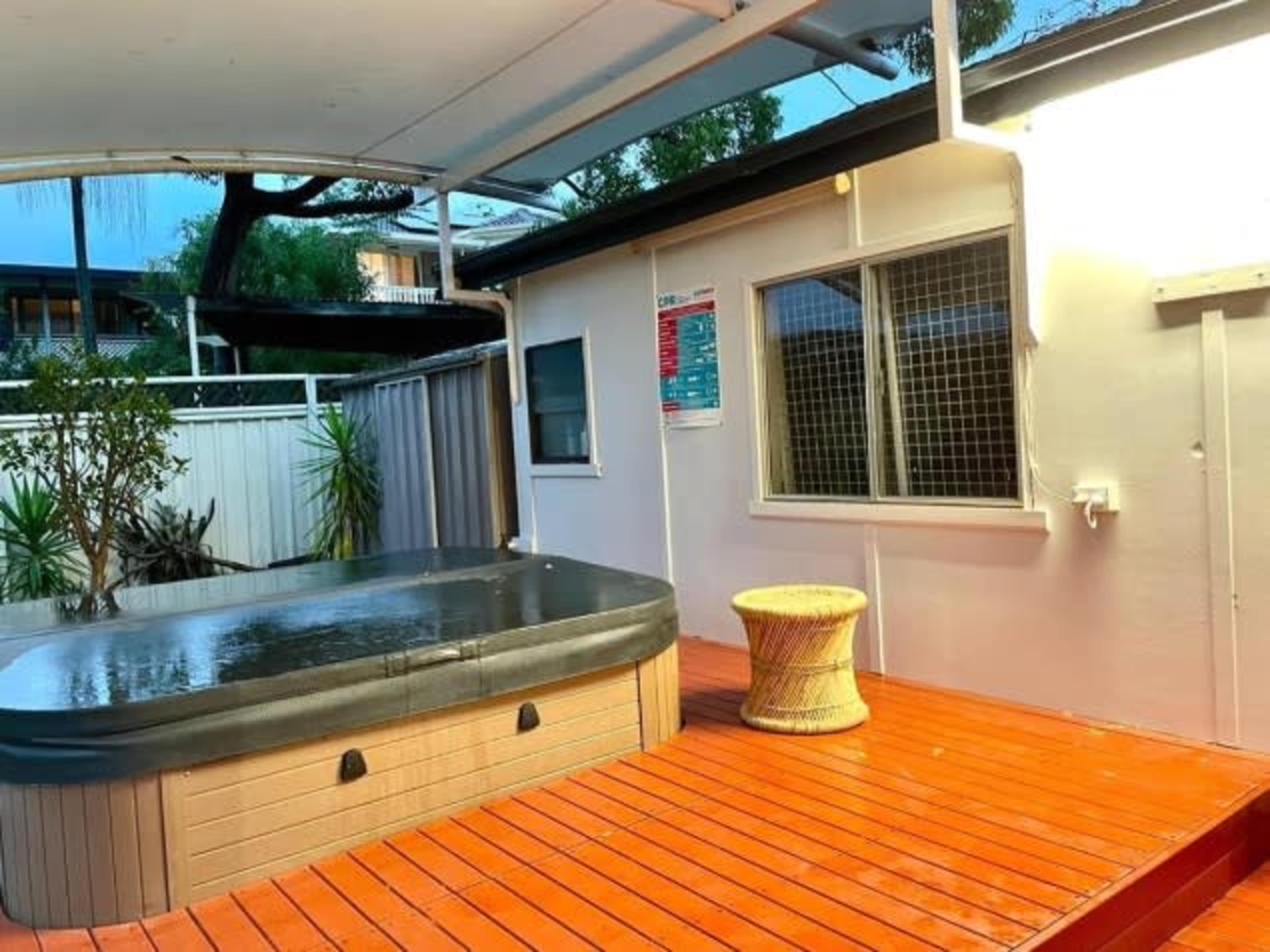 A wooden deck area featuring a hot tub and a wicker stool, surrounded by plants and a building with windows.