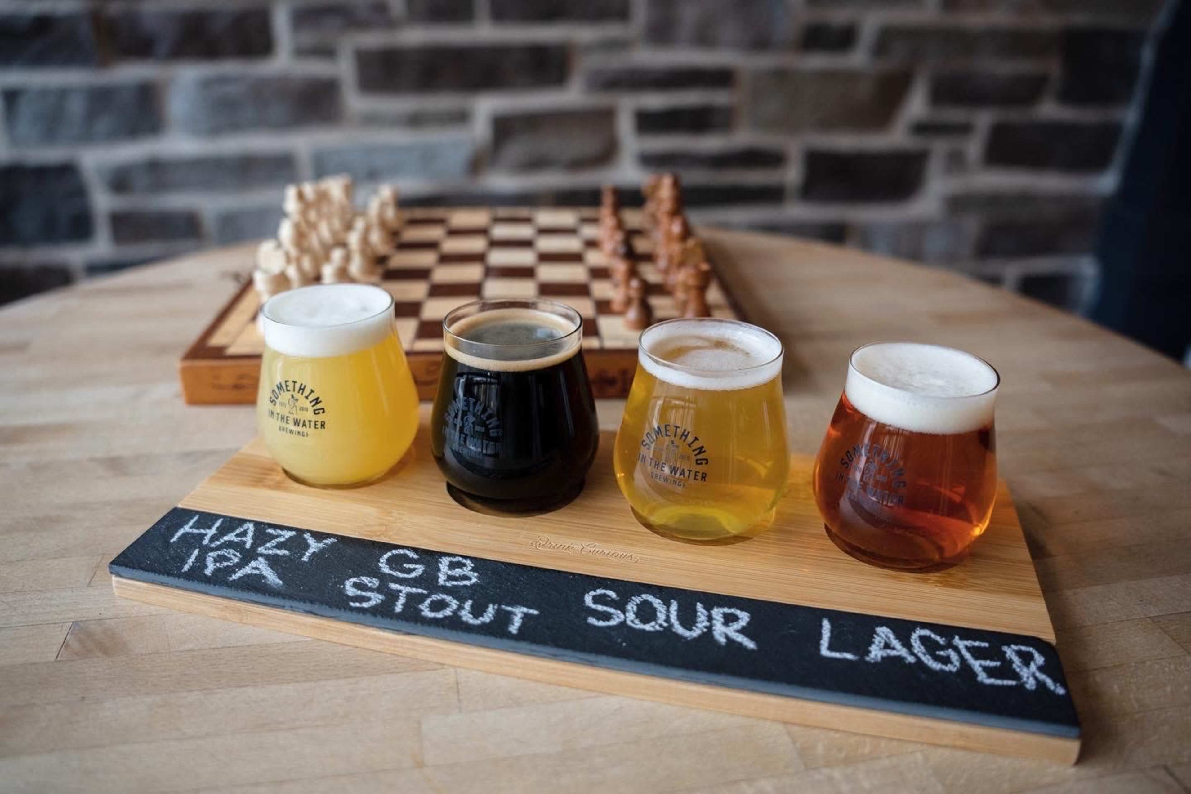 A wooden flight of four craft beers is displayed next to a chessboard on a stone wall background.