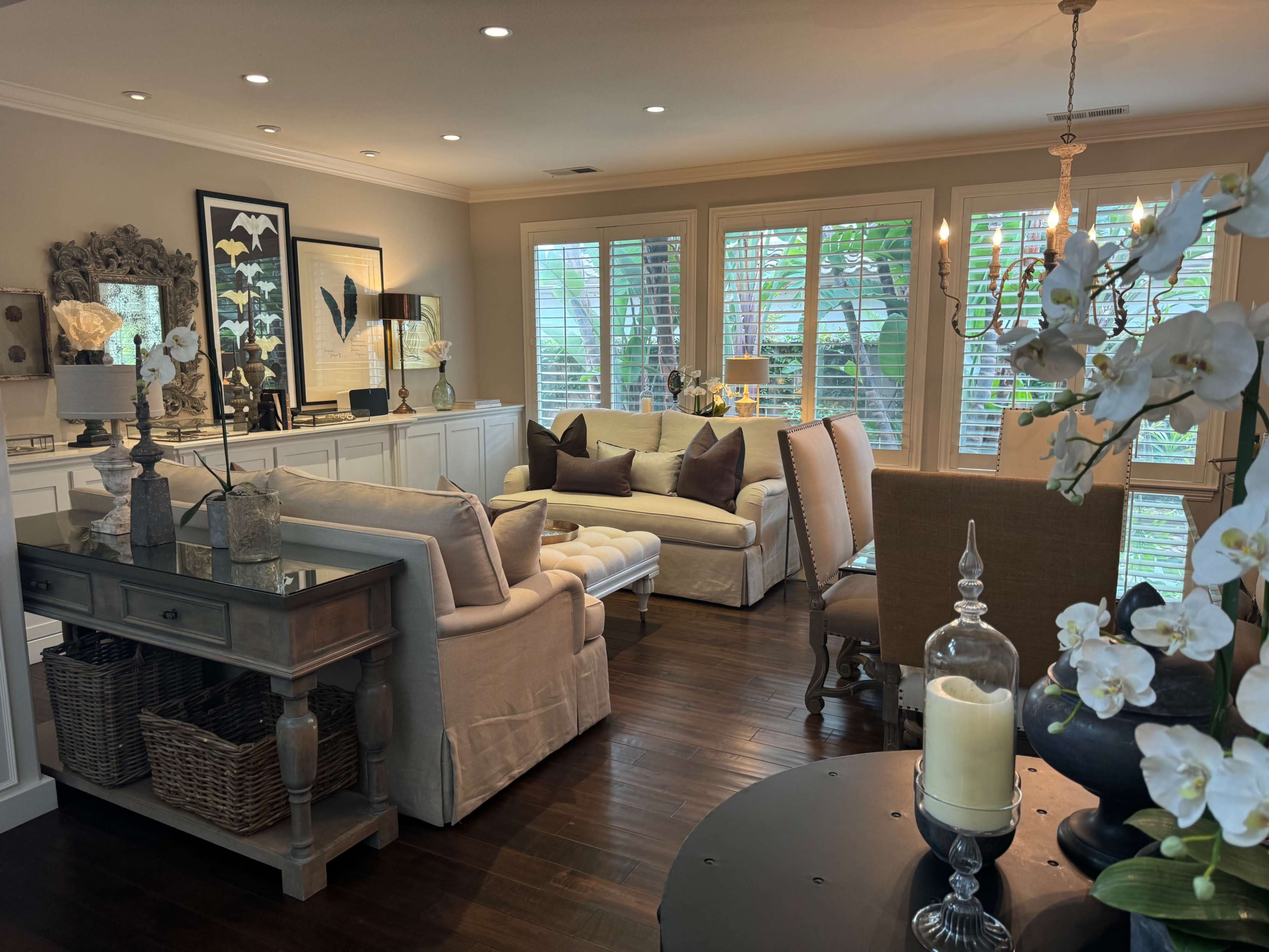 A well-furnished living room featuring a light-colored sofa, a wooden coffee table, and large windows letting in natural light, alongside decorative plants and artwork.