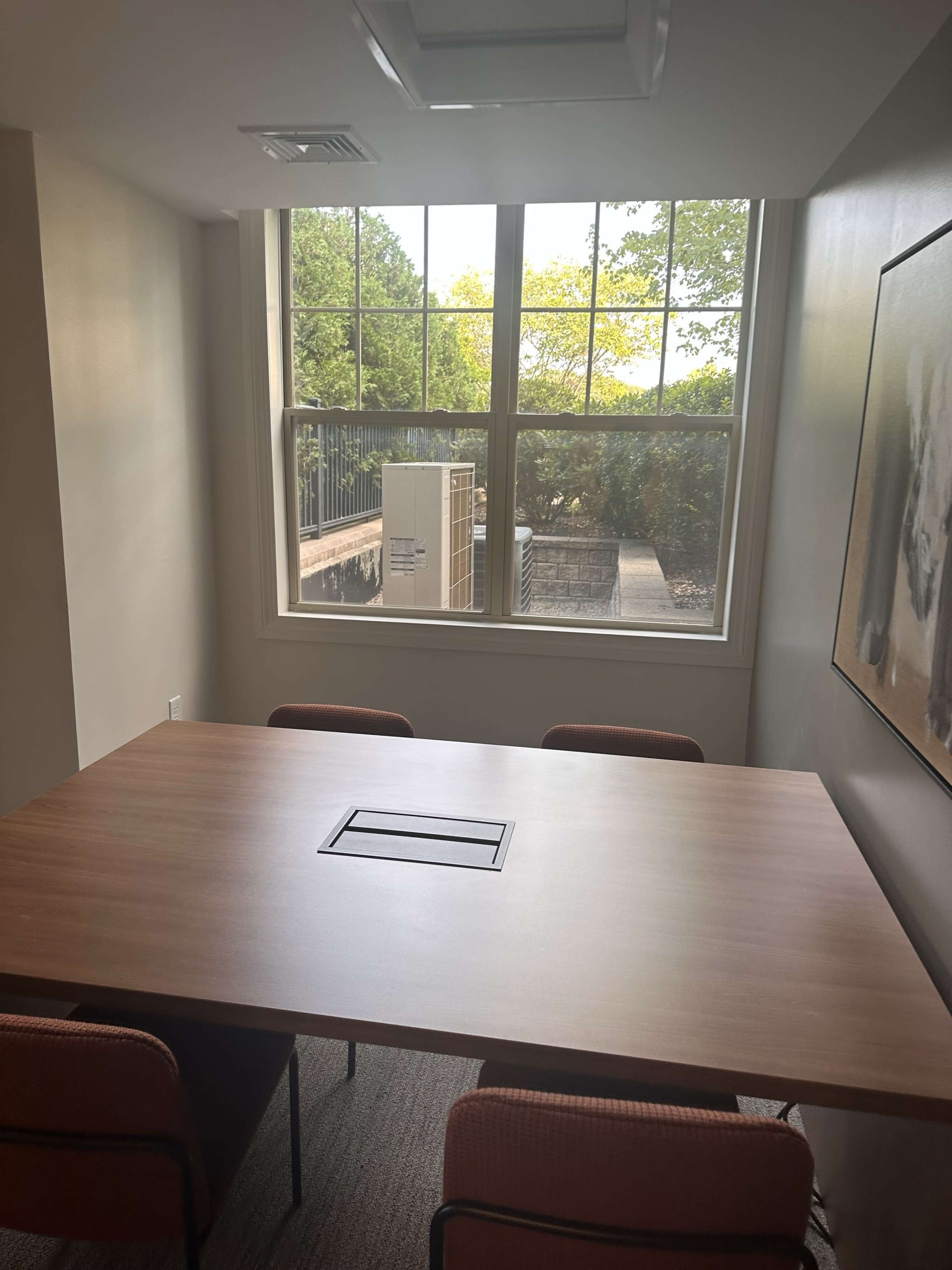 A small meeting room with a wooden table and four chairs, featuring a large window that overlooks an outdoor area.