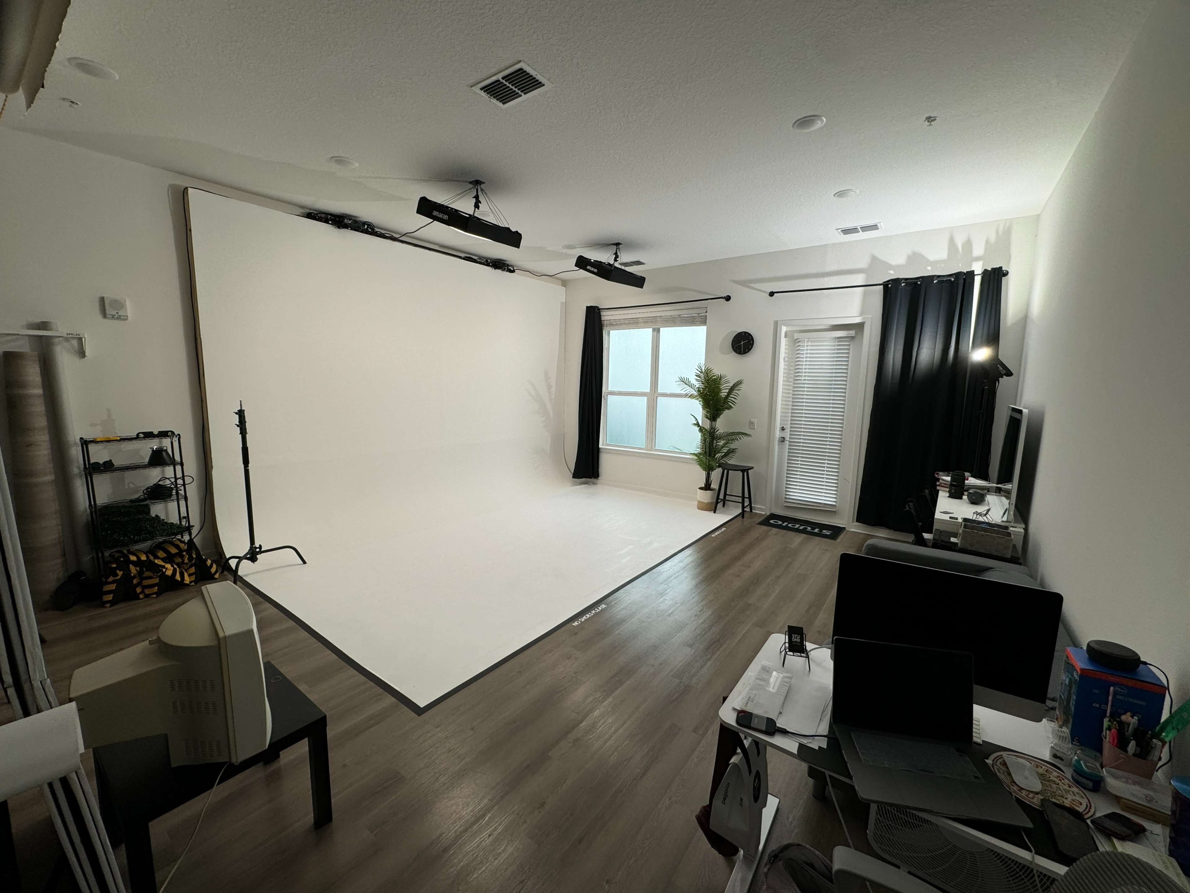 The image shows a bright, minimalist photography studio with a white backdrop, windows, and a desk setup.
