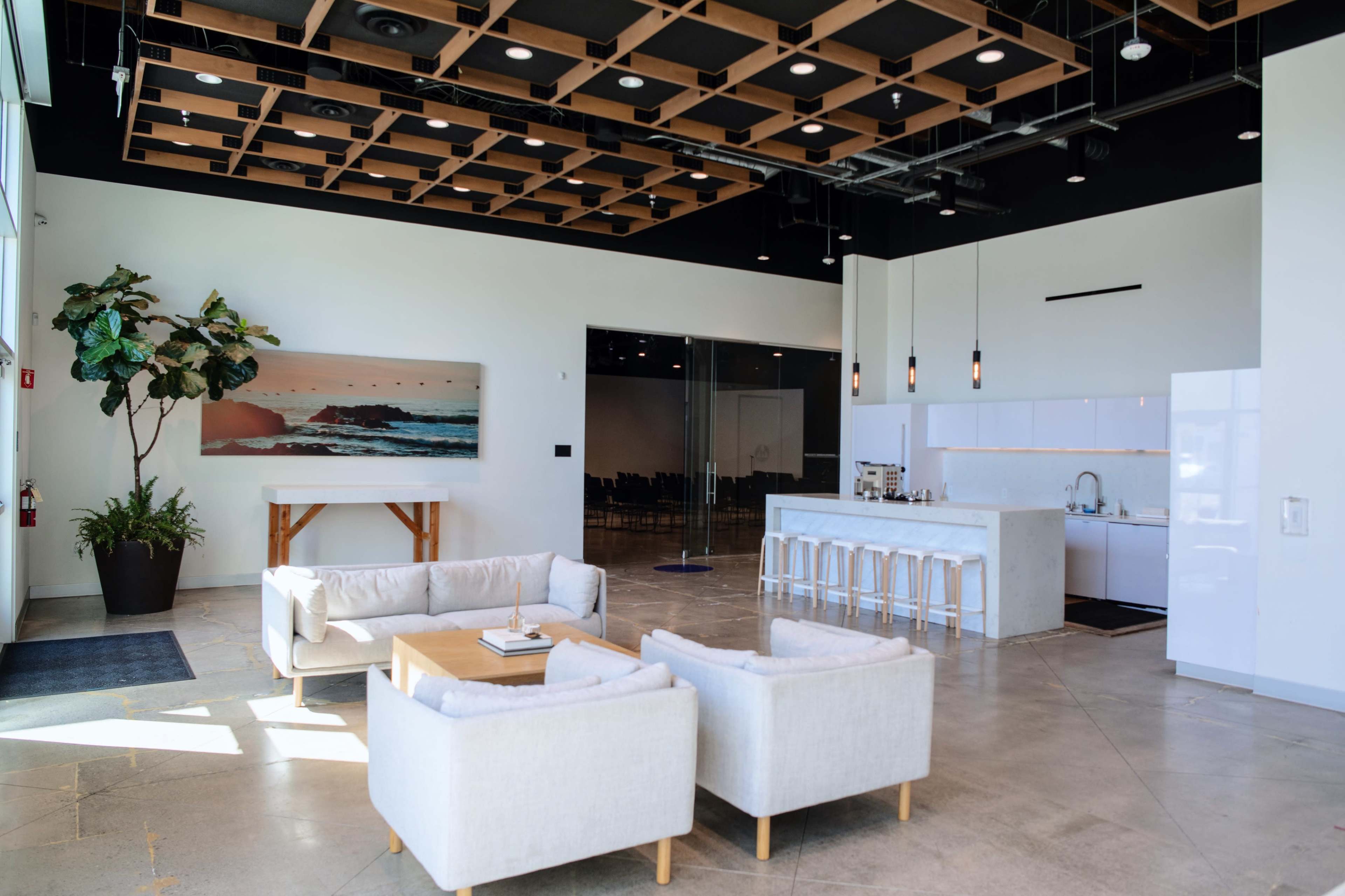 The image shows a modern lobby area featuring a seating arrangement with light-colored couches, a kitchen space with barstools, and large windows allowing natural light to enter.