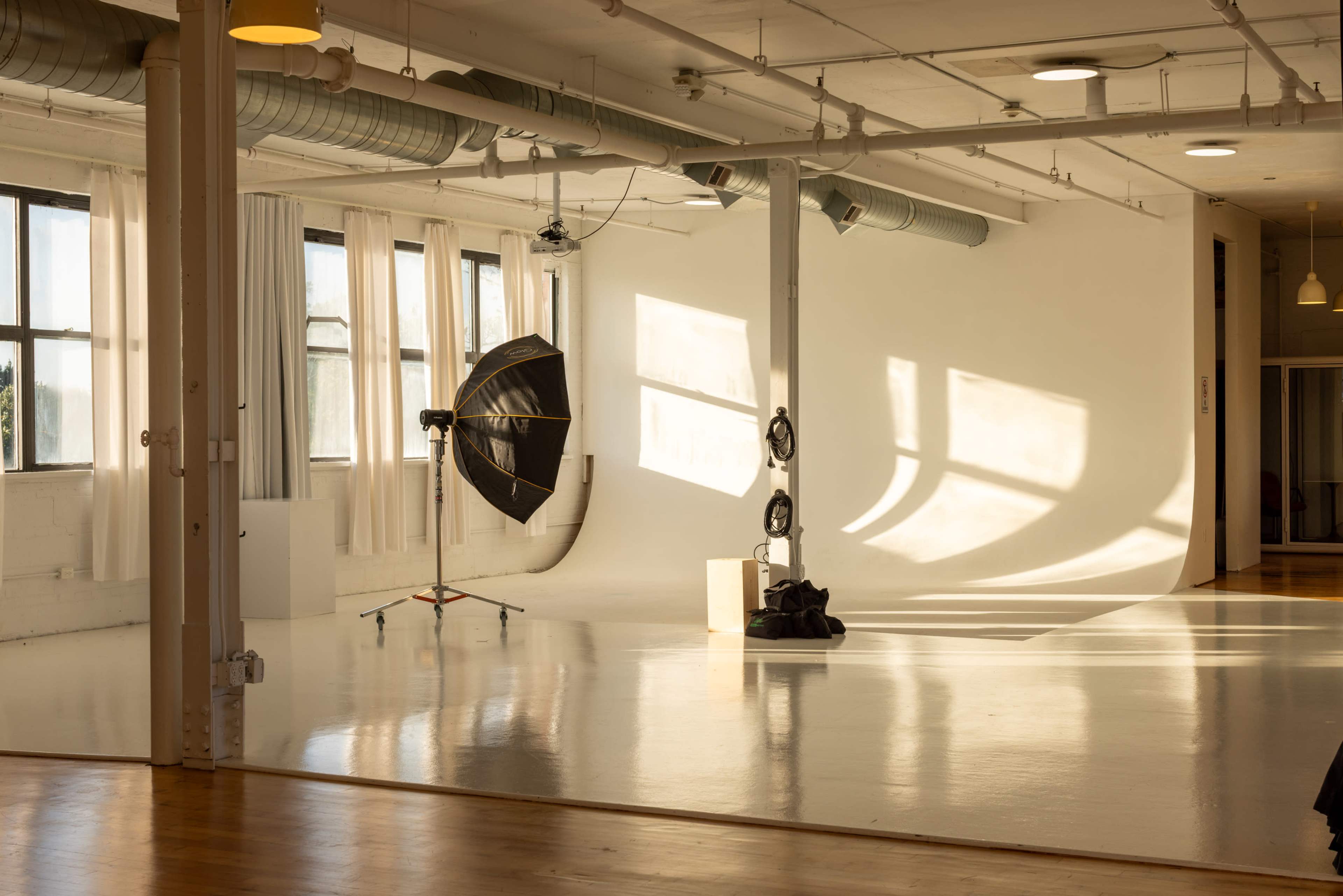 The image shows a spacious photography studio with large windows, a softbox light on a stand, and warm sunlight casting shadows on the white walls.