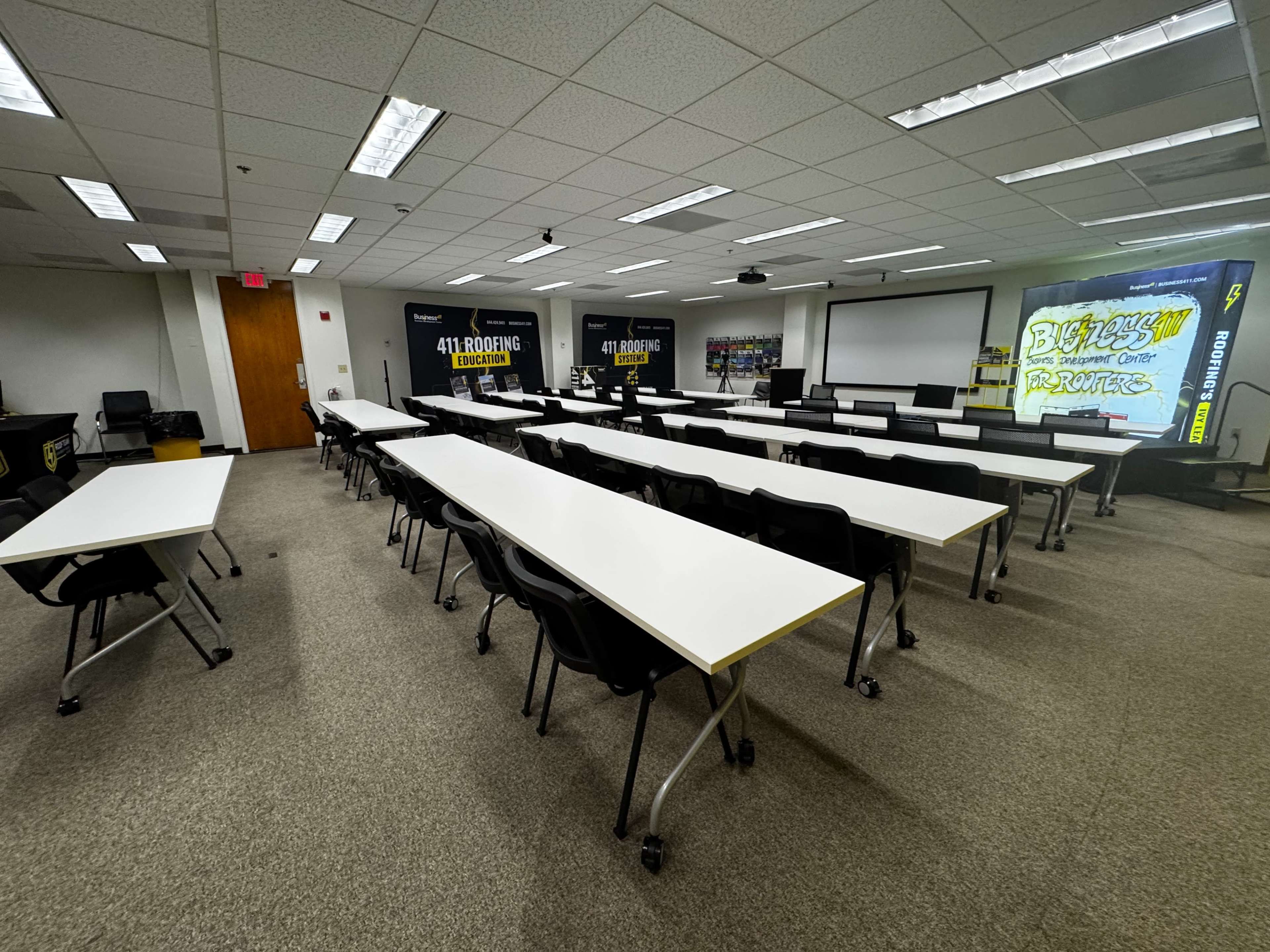 The image shows a spacious classroom or conference room arranged with multiple long tables and black chairs, featuring large screens on the walls.