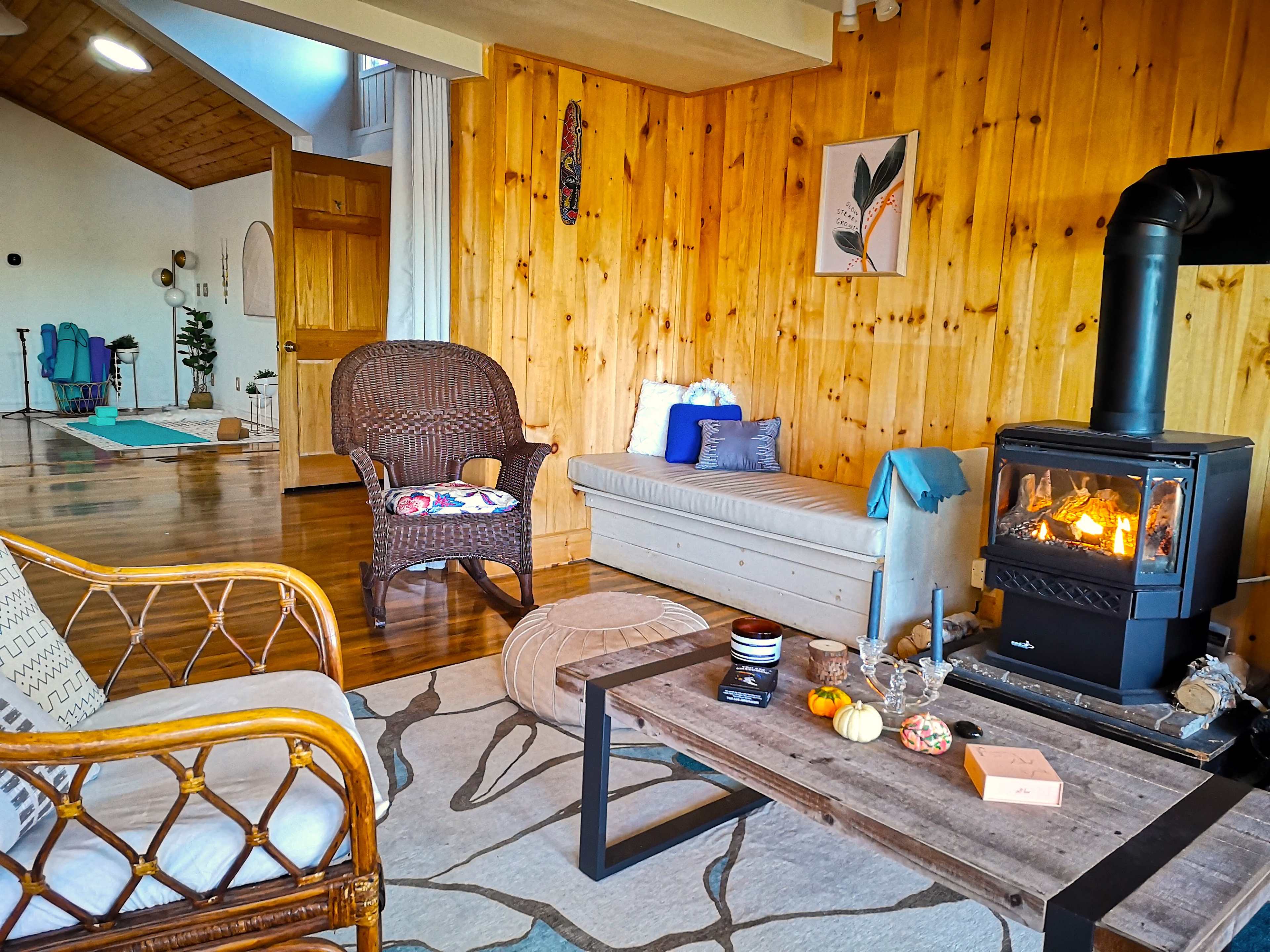 A cozy living room features a wood stove, wooden walls, and a combination of seating areas with a coffee table adorned with decorative items.