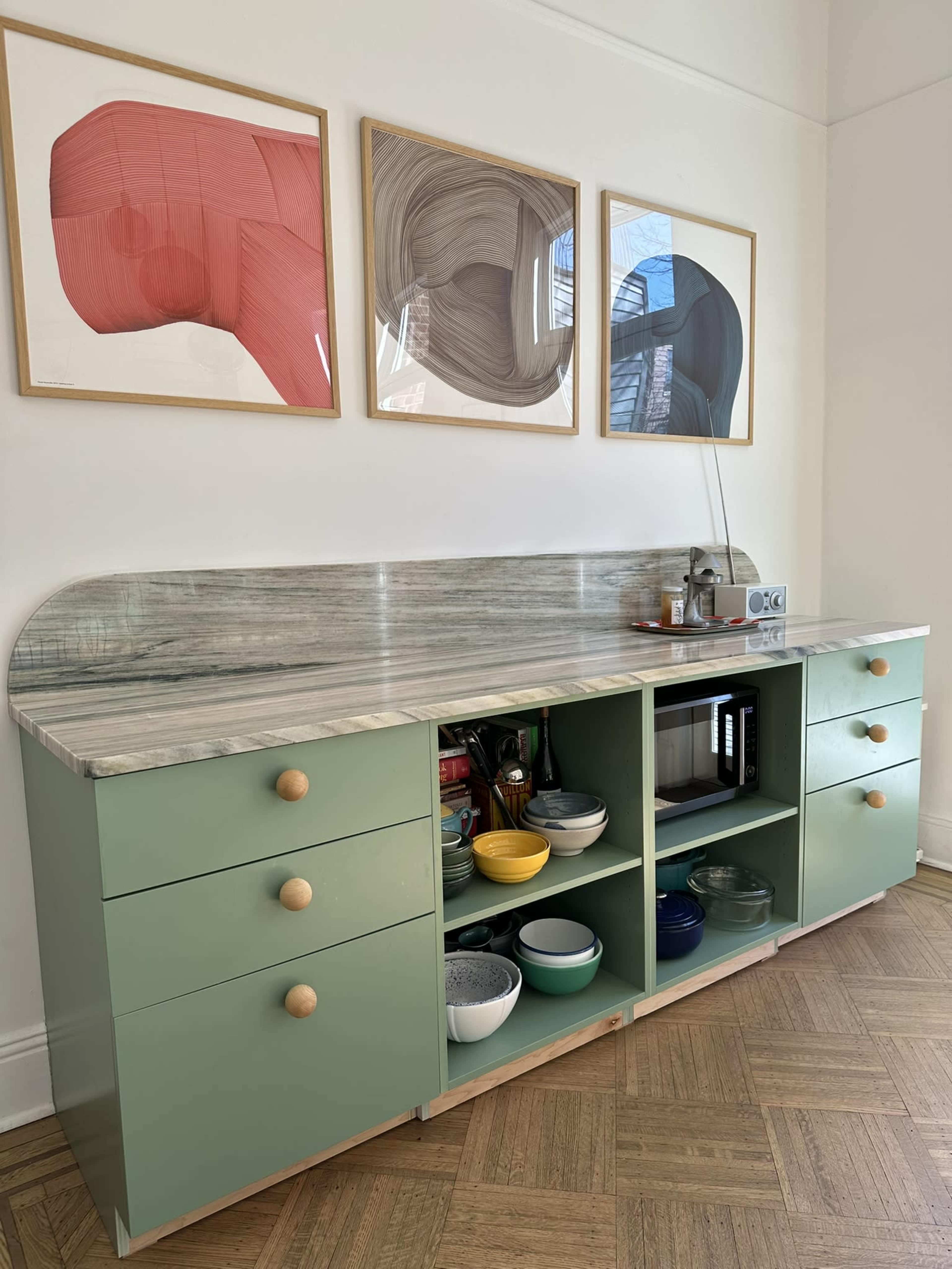 A modern kitchen cabinet painted green features a marble countertop and is adorned with artwork above, showcasing various dishes organized within the cabinet.