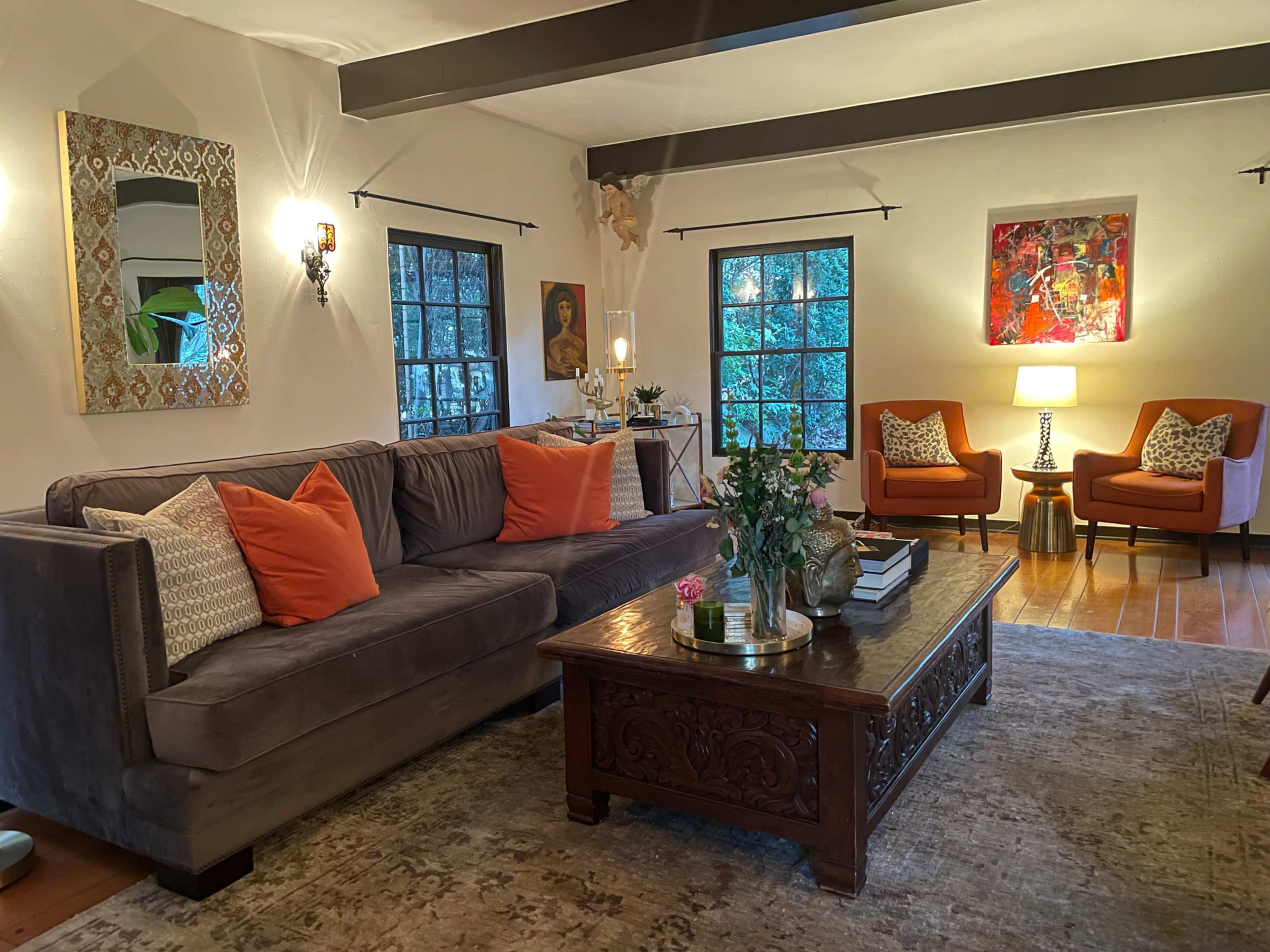 The living room features a gray sofa with orange accent pillows, two orange armchairs, a wooden coffee table, and large windows allowing natural light to enter.