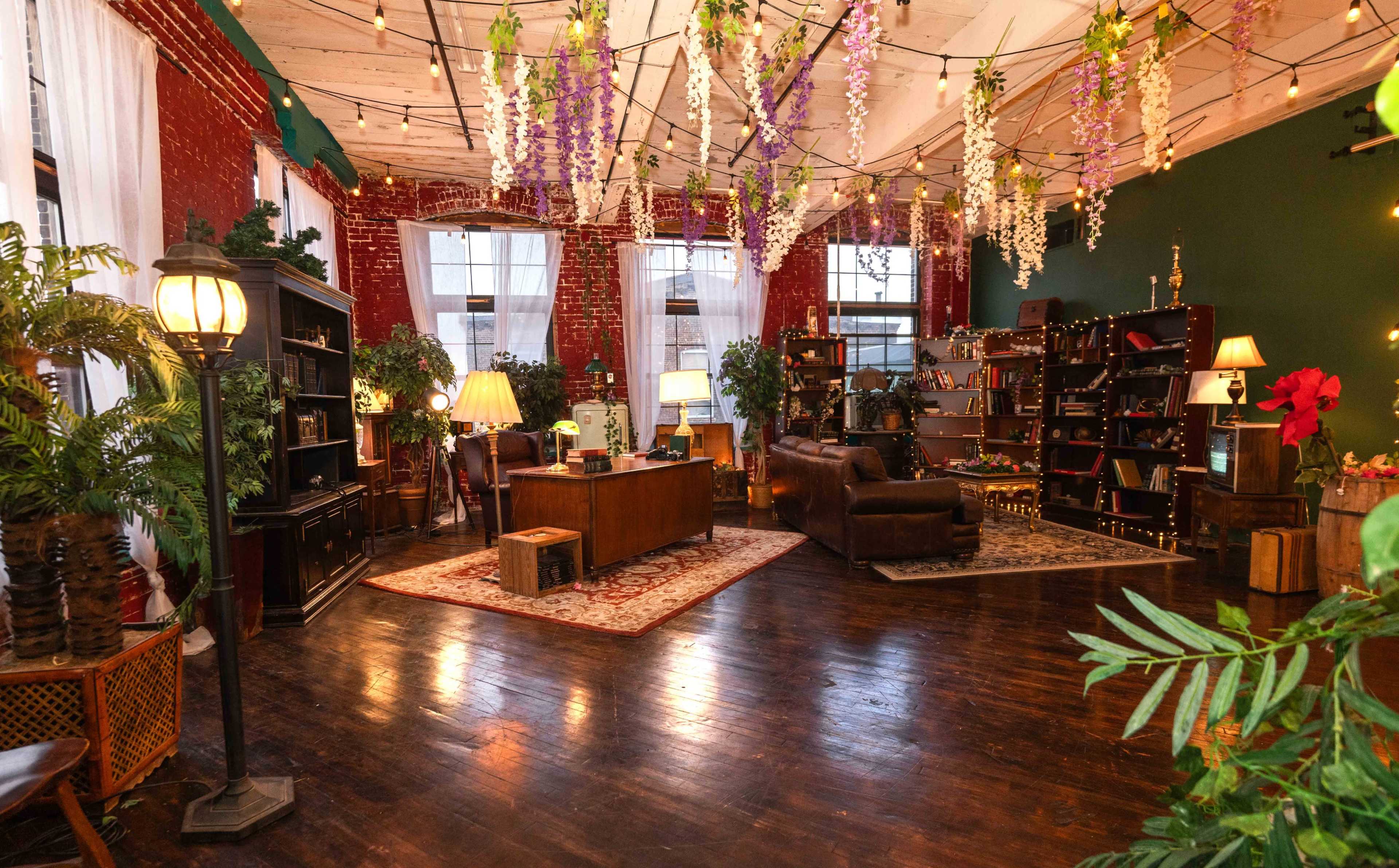 The image shows a brightly lit room with red brick walls, wooden floors, and various plants, featuring a desk, bookshelves, and decorated with hanging flowers and string lights.