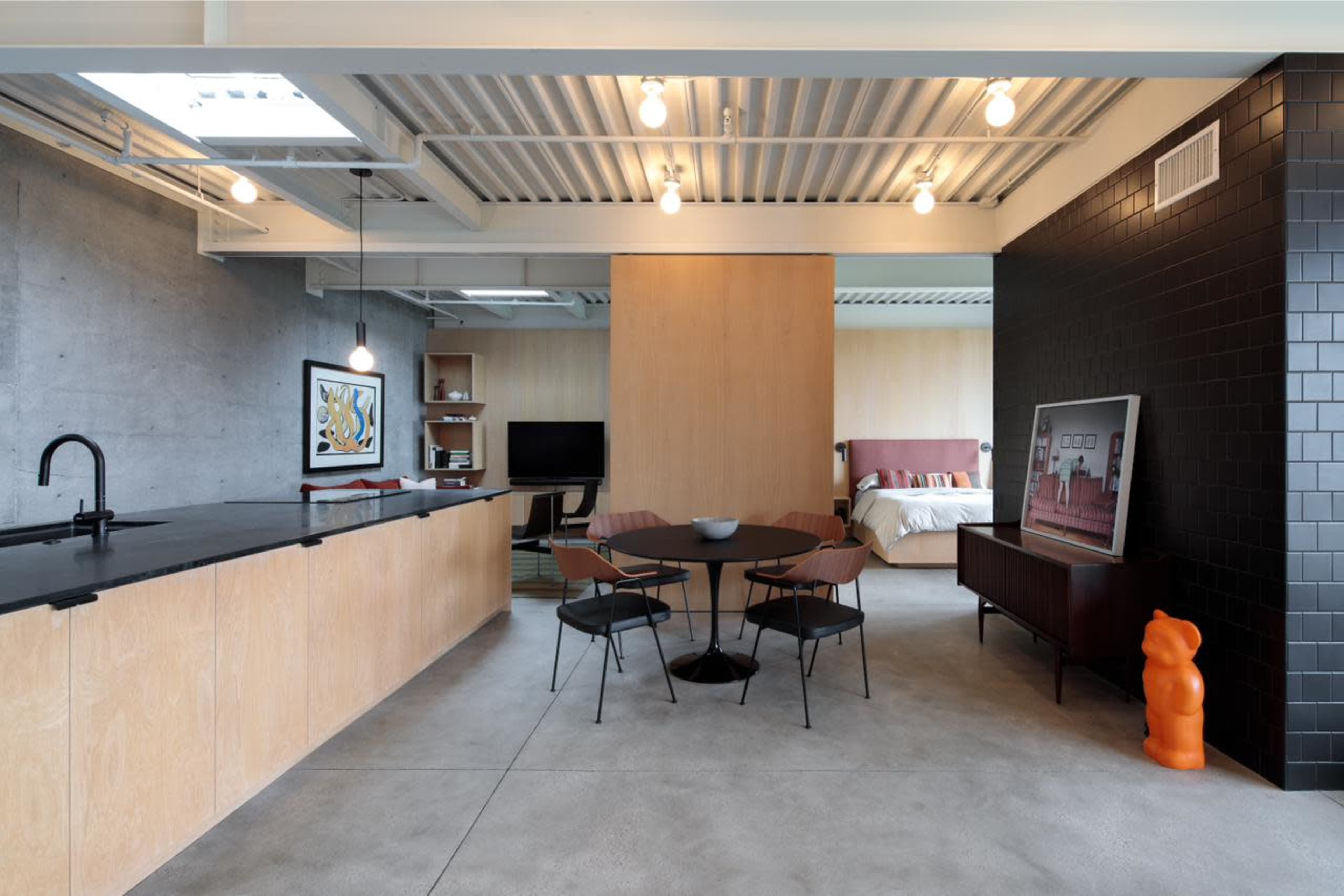 The image depicts a modern, open-concept living space featuring a kitchen area with a wooden bar, a dining table with chairs, and a bedroom in the background separated by a wall.