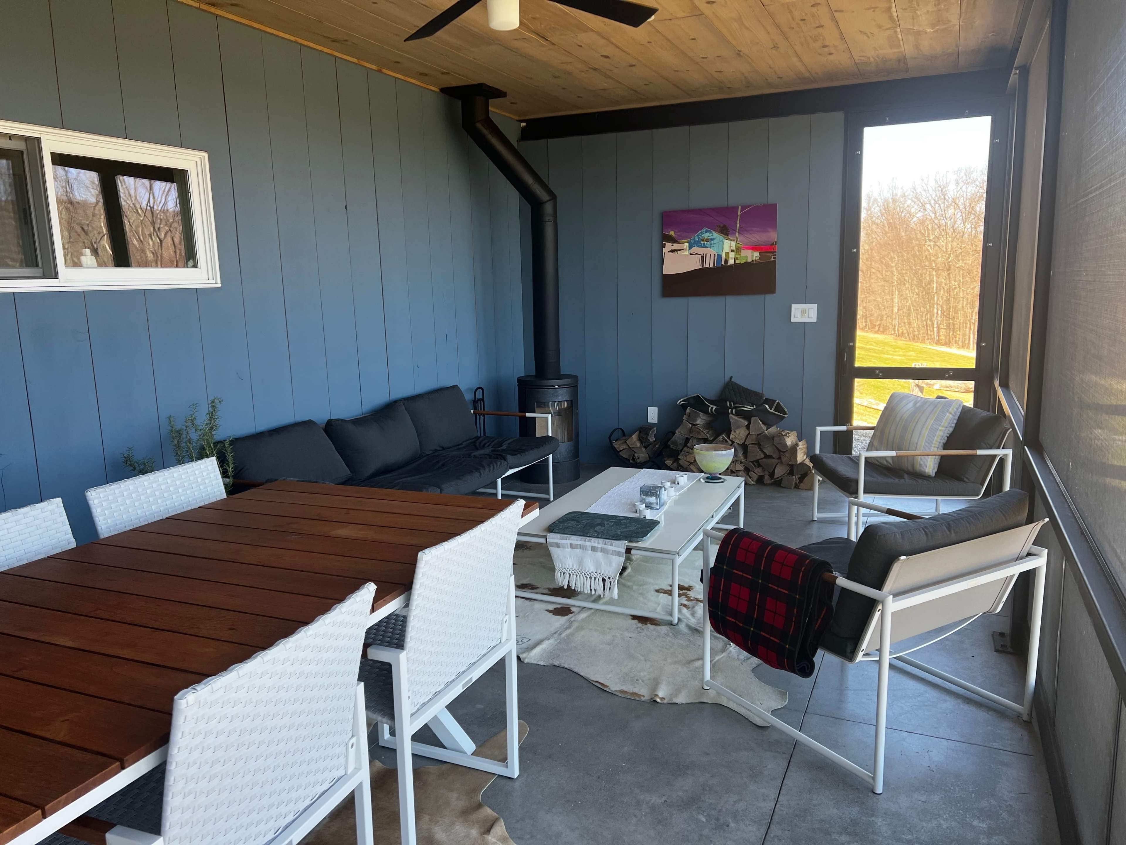 A covered patio features a dining table, seating area with a sectional sofa and accent chairs, a wood stove, and a decorative wall art piece.