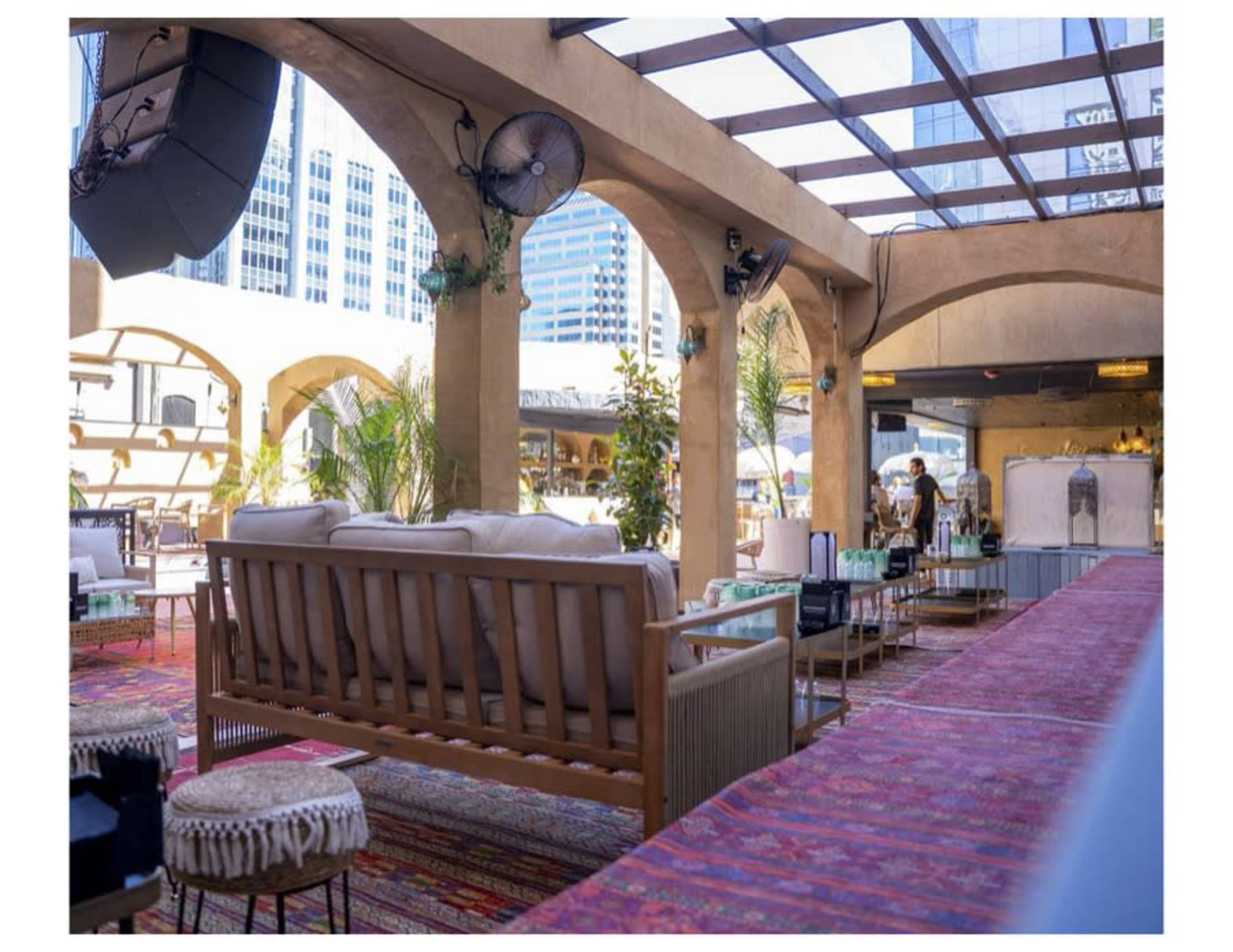 The image depicts a modern rooftop lounge area featuring wooden furniture, potted plants, and a view of city buildings in the background.