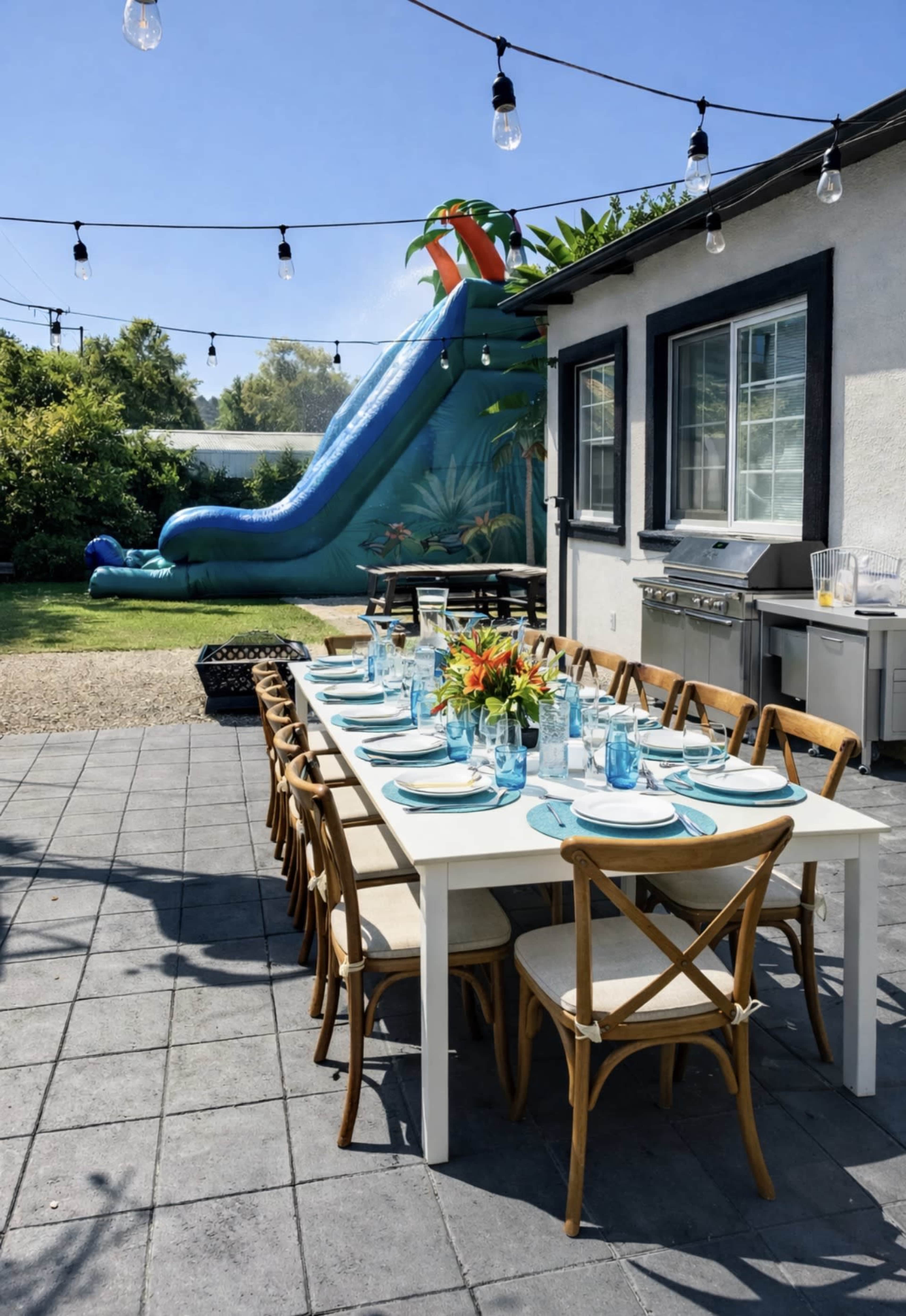 A long outdoor dining table is set with blue glassware and place settings, while a large waterslide is positioned in the background against a sunny yard.