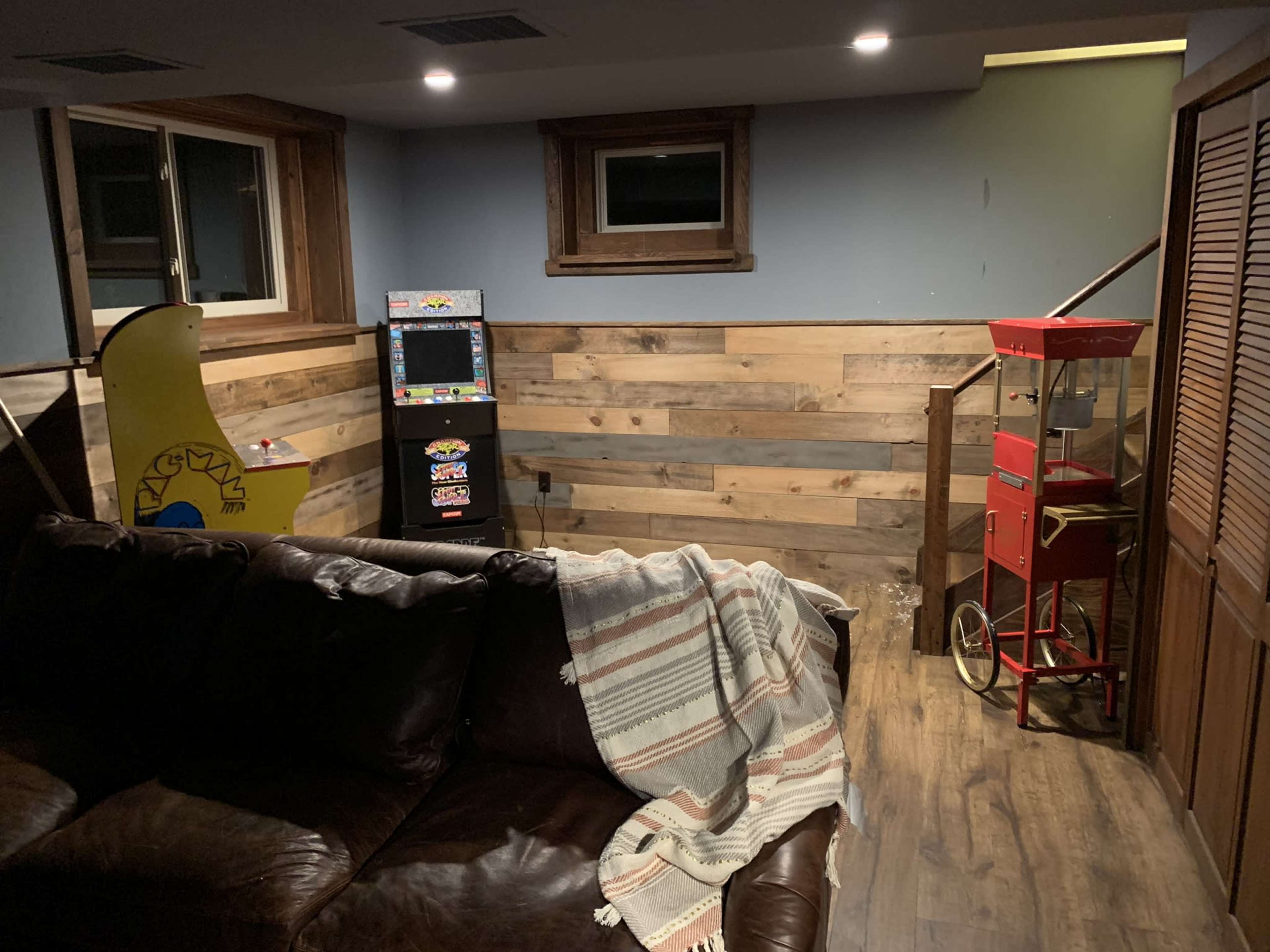 A basement room with a brown leather couch draped with a blanket, a retro arcade game, and a vintage popcorn machine near a staircase.