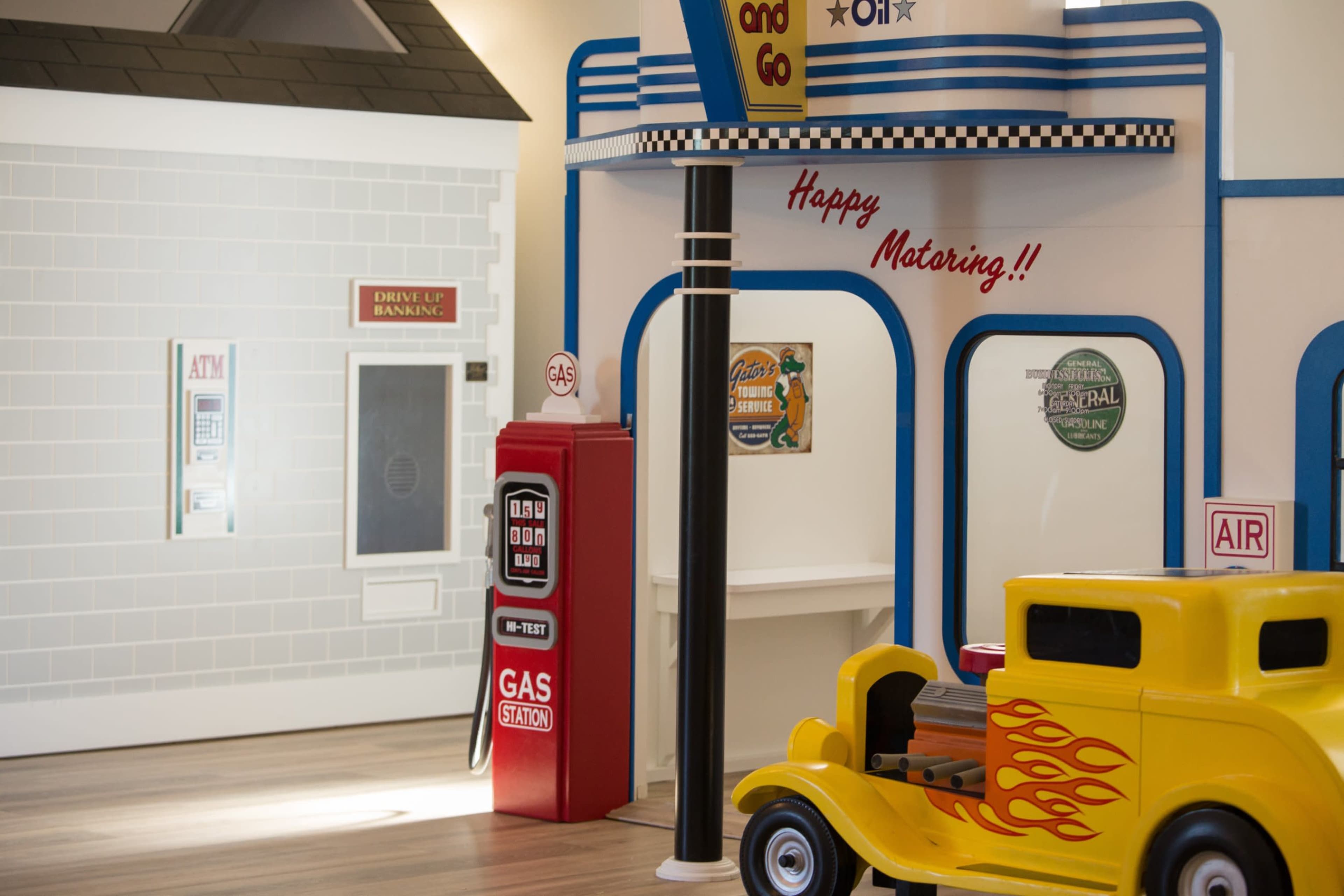 The image shows a vibrant children's play area designed to resemble a gas station, complete with a red gas pump, a toy car, and a wall with a drive-thru sign.