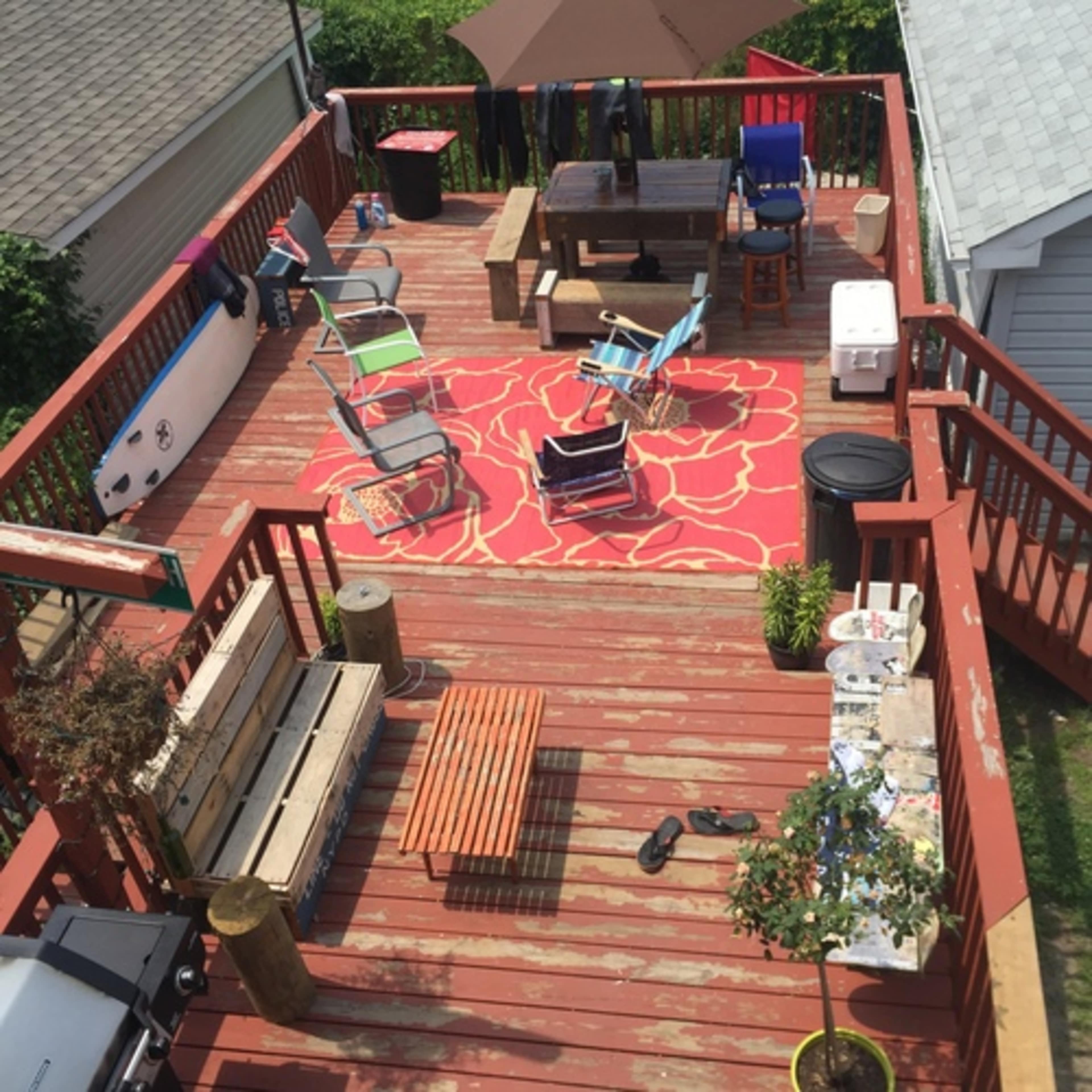 A wooden deck features various chairs, a dining table, a cooler, and decorative elements, all set on a floral area rug.