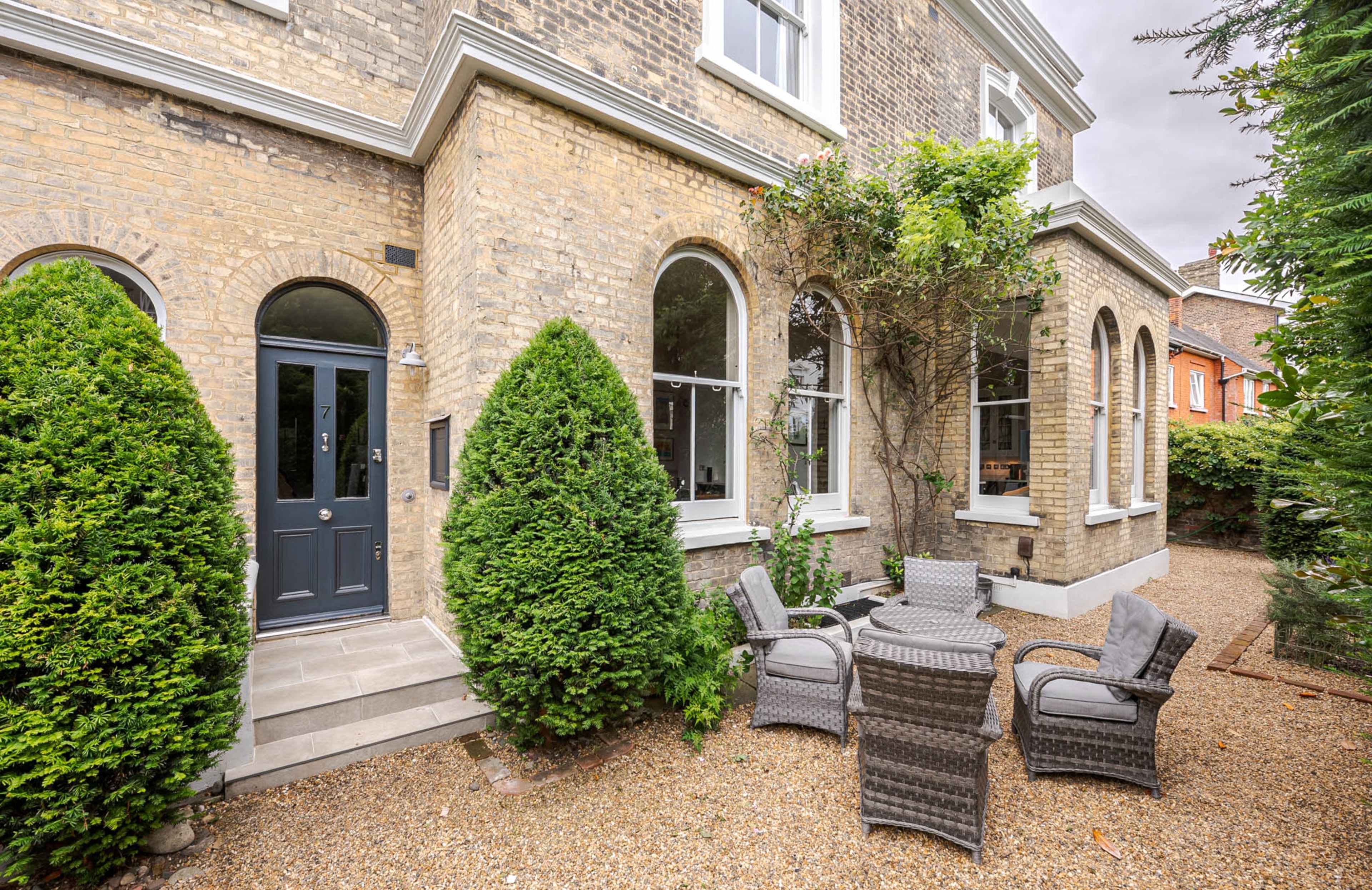 The image shows a brick house entrance with a dark door flanked by arching windows, surrounded by neatly trimmed shrubs and a small gravel patio with wicker furniture.