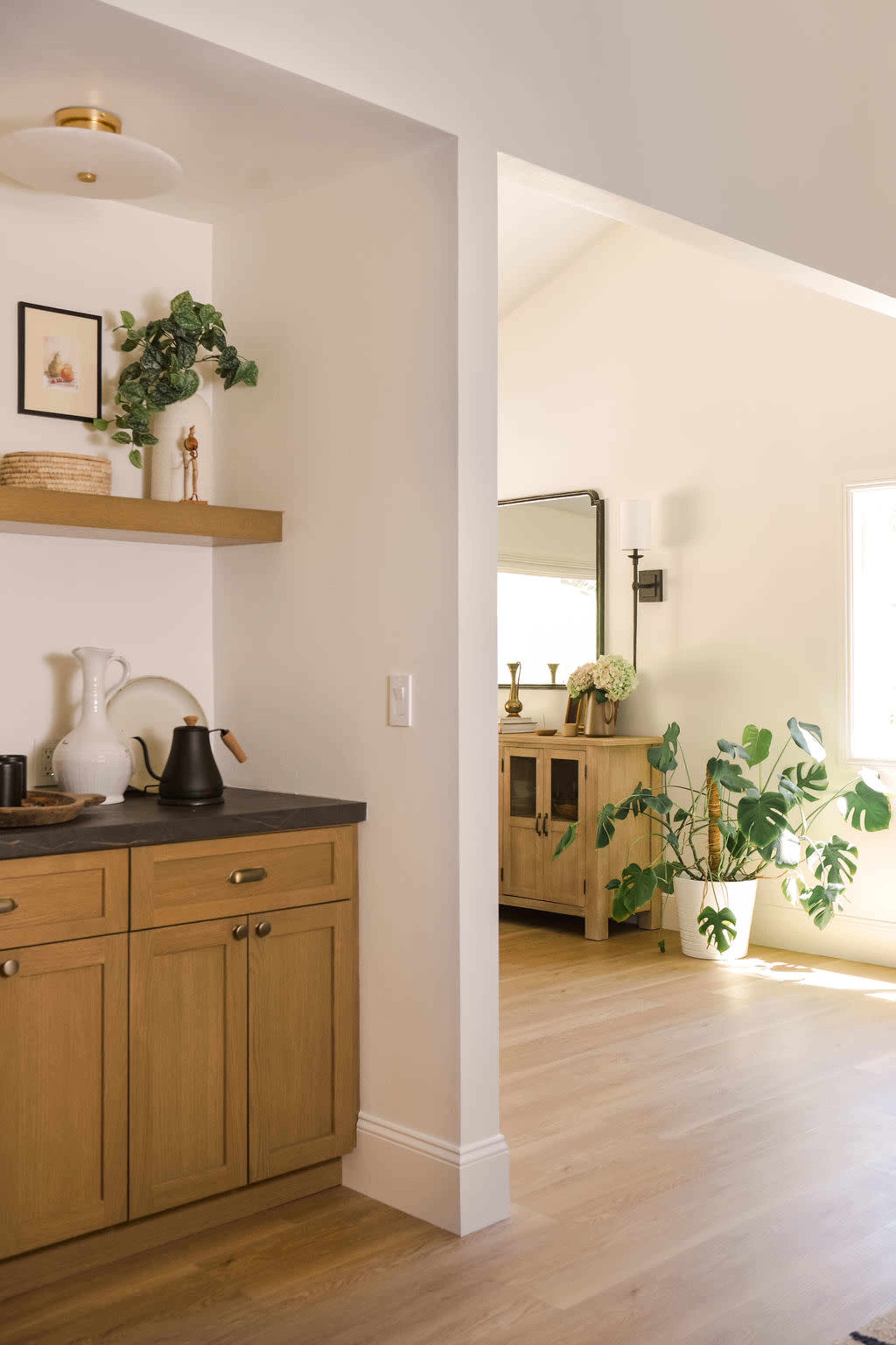 A modern kitchen area features light wood cabinetry, a black countertop with a kettle, and plants, visible through an open doorway leading to a brightly lit room.