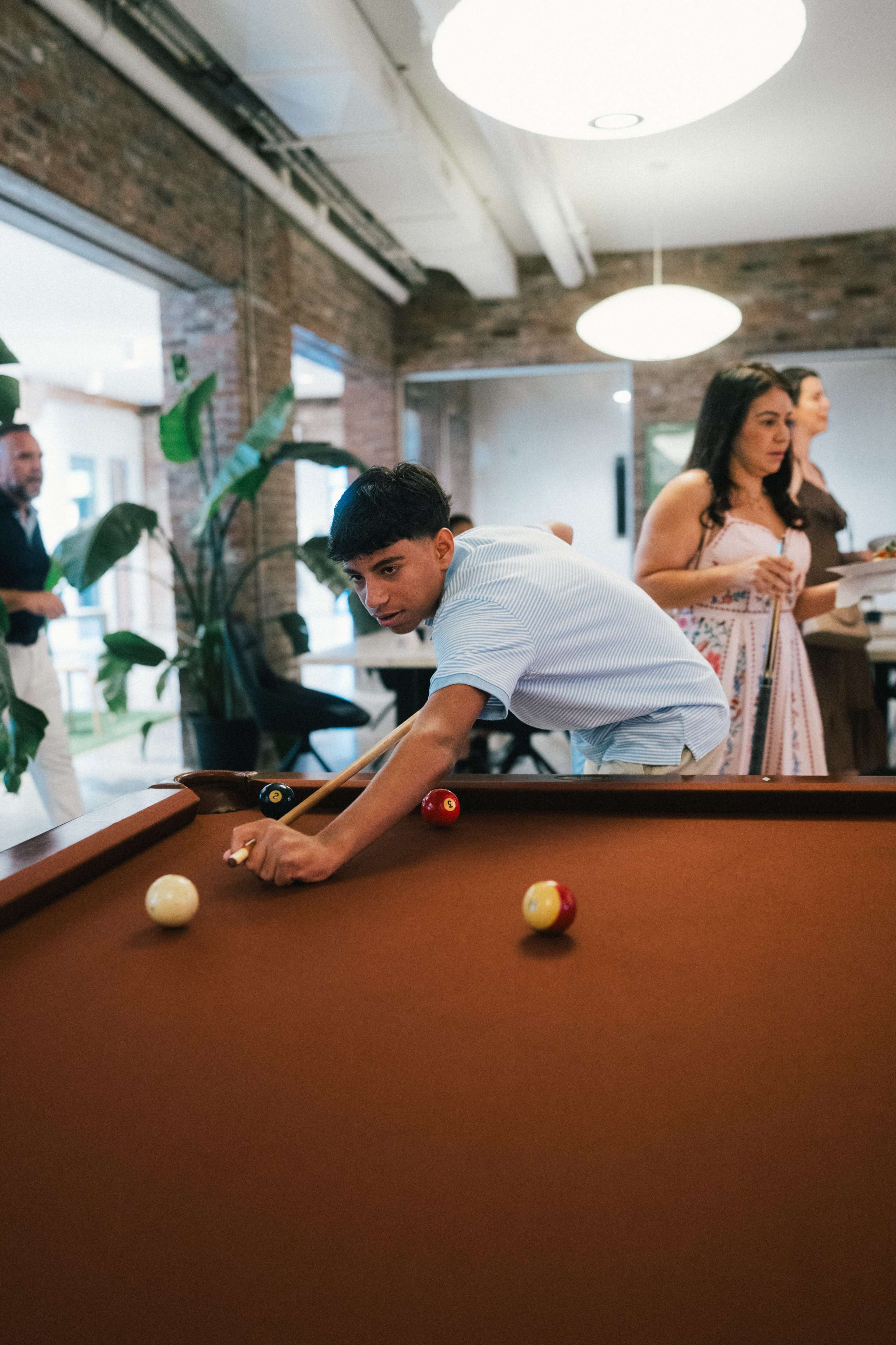 A young man leans over a pool table, aiming to strike a ball while others socialize in the background.