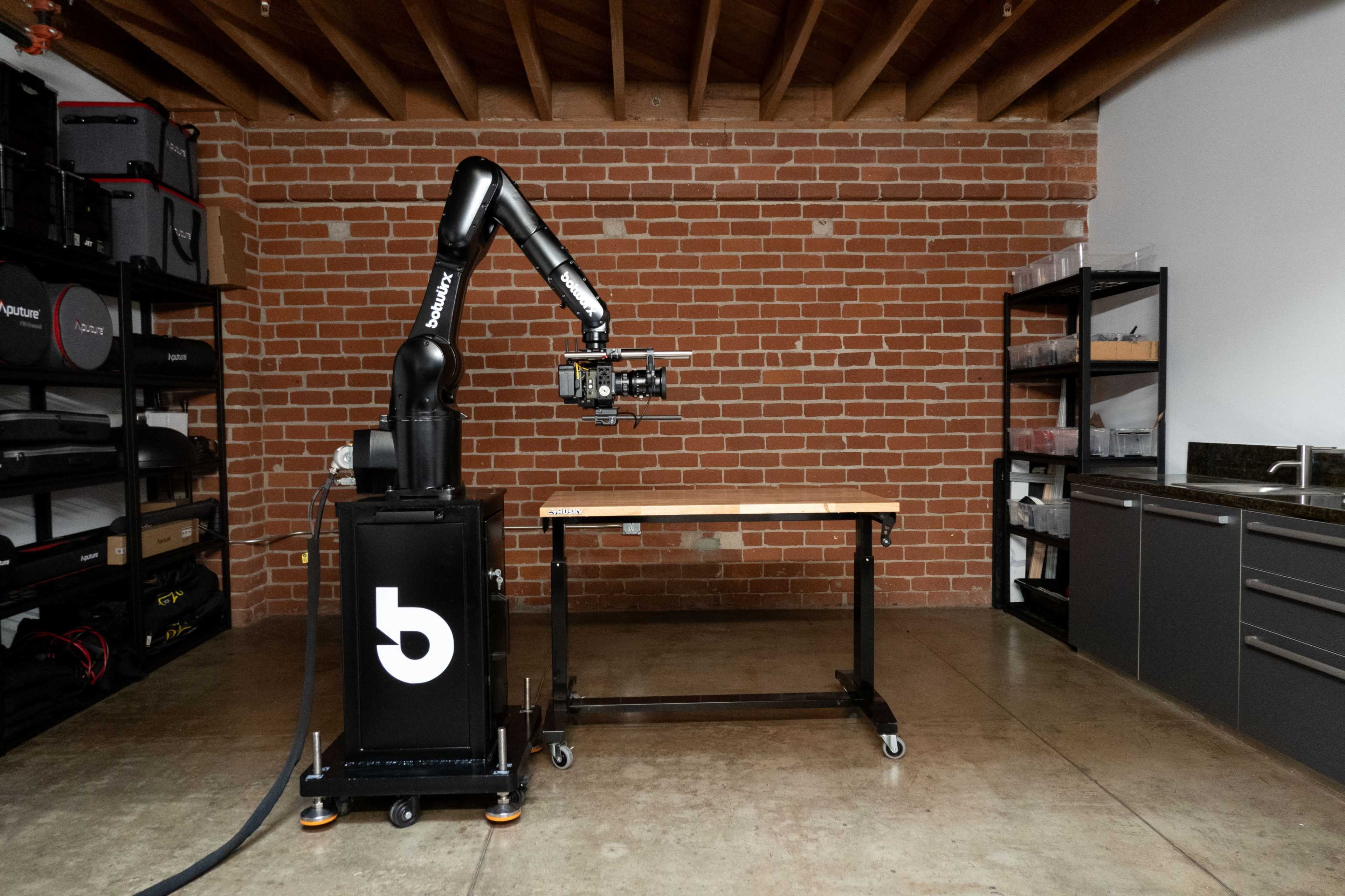 A robotic arm is positioned next to a wooden table in a workshop with exposed brick walls and functional storage cabinets.
