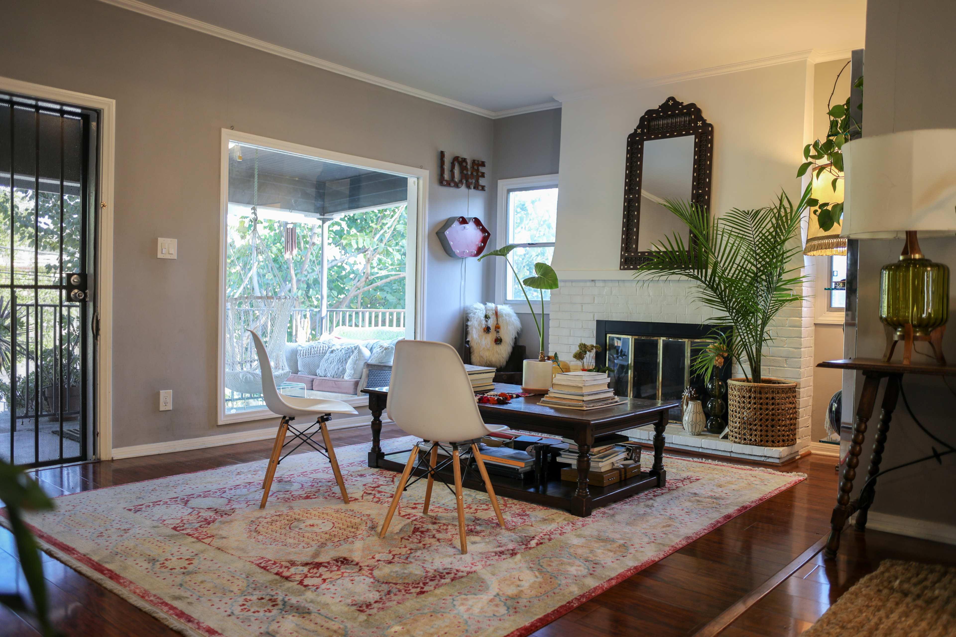 The scene depicts a cozy living room with a coffee table, two white chairs, a fireplace, and large windows that allow natural light to enter.