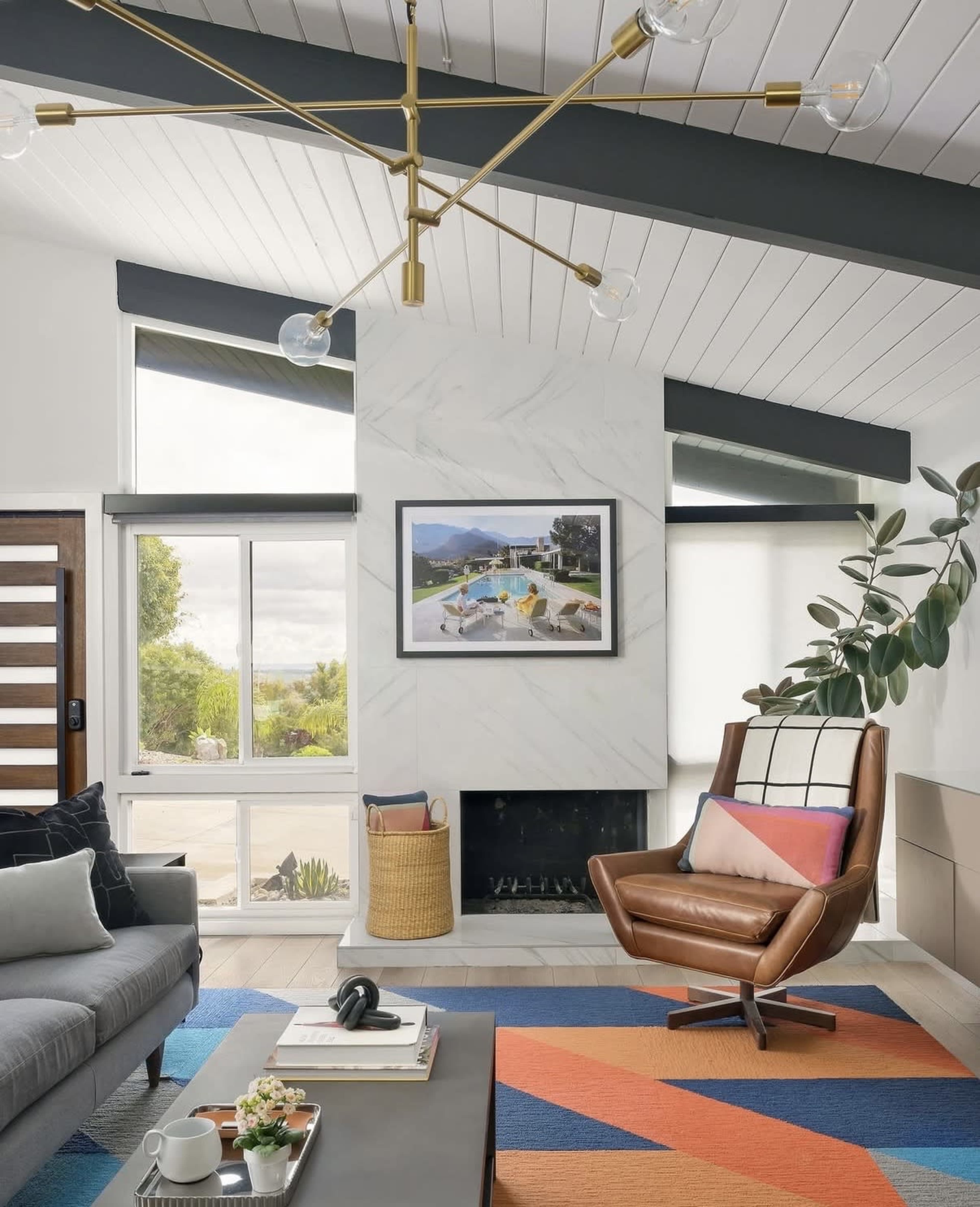 A modern living room features a gray sofa, a brown swivel chair, a marble fireplace, and a colorful area rug under a geometric light fixture.