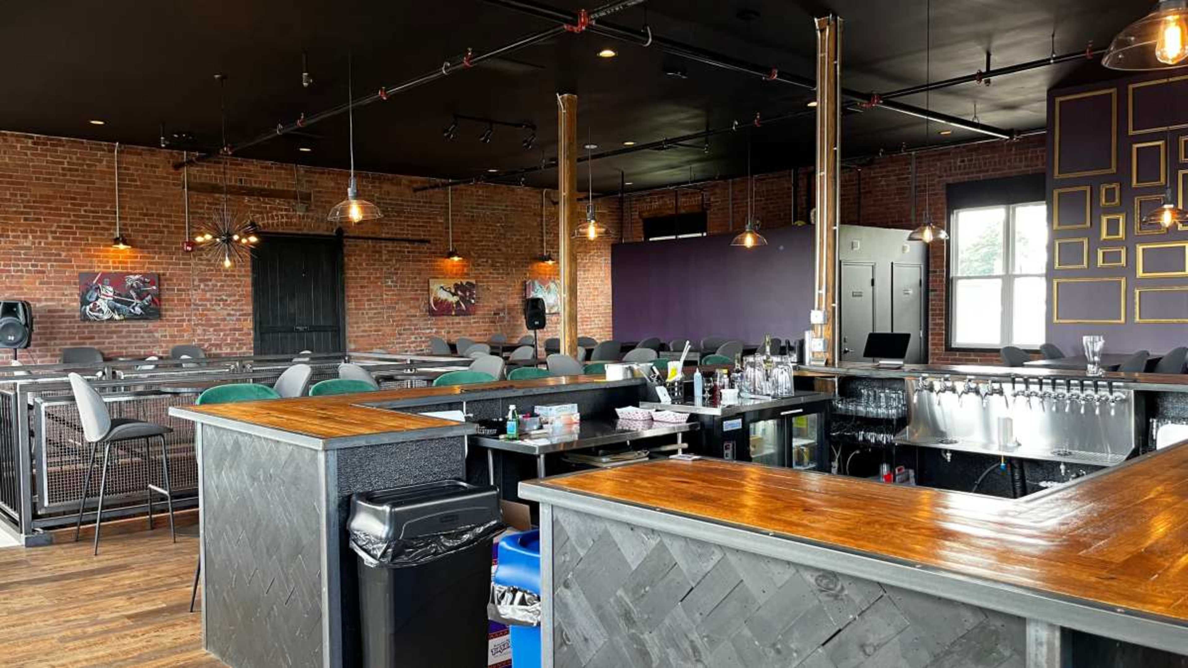 A modern bar interior with exposed brick walls, a wooden bar counter, and rows of seating arranged in a spacious layout.