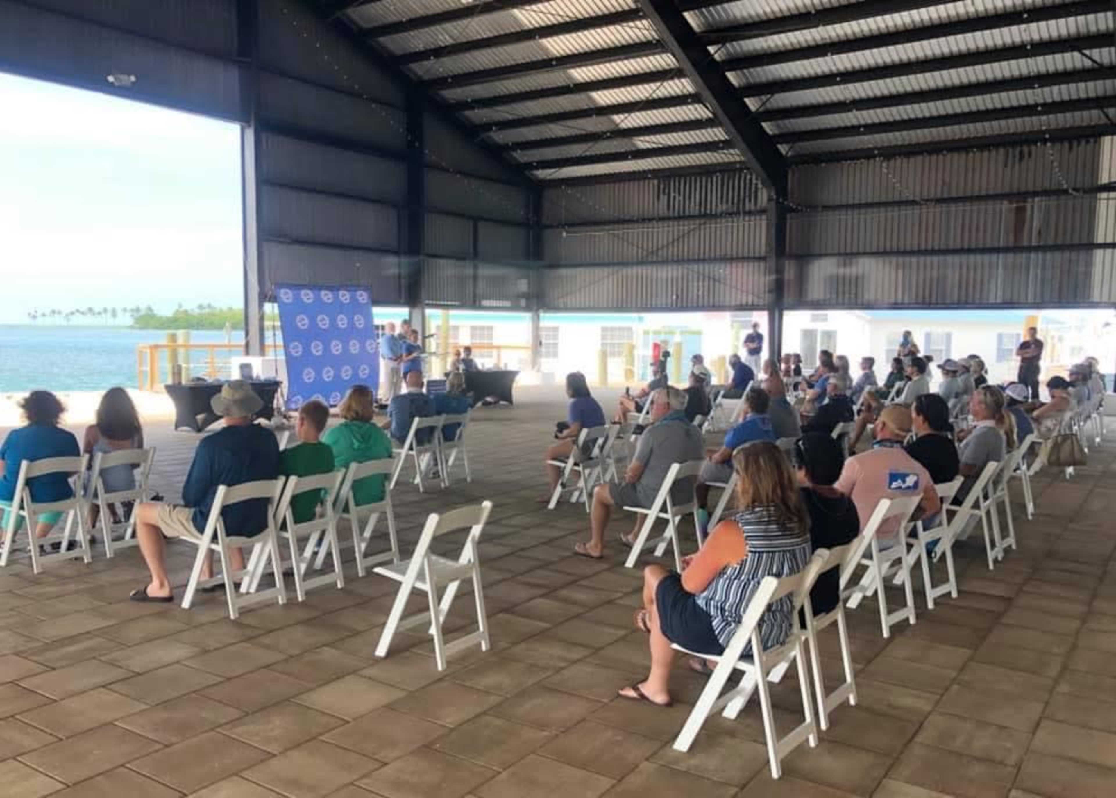 A crowd of people sits in folding chairs under a large metal structure, listening to a speaker in front of a branded backdrop.