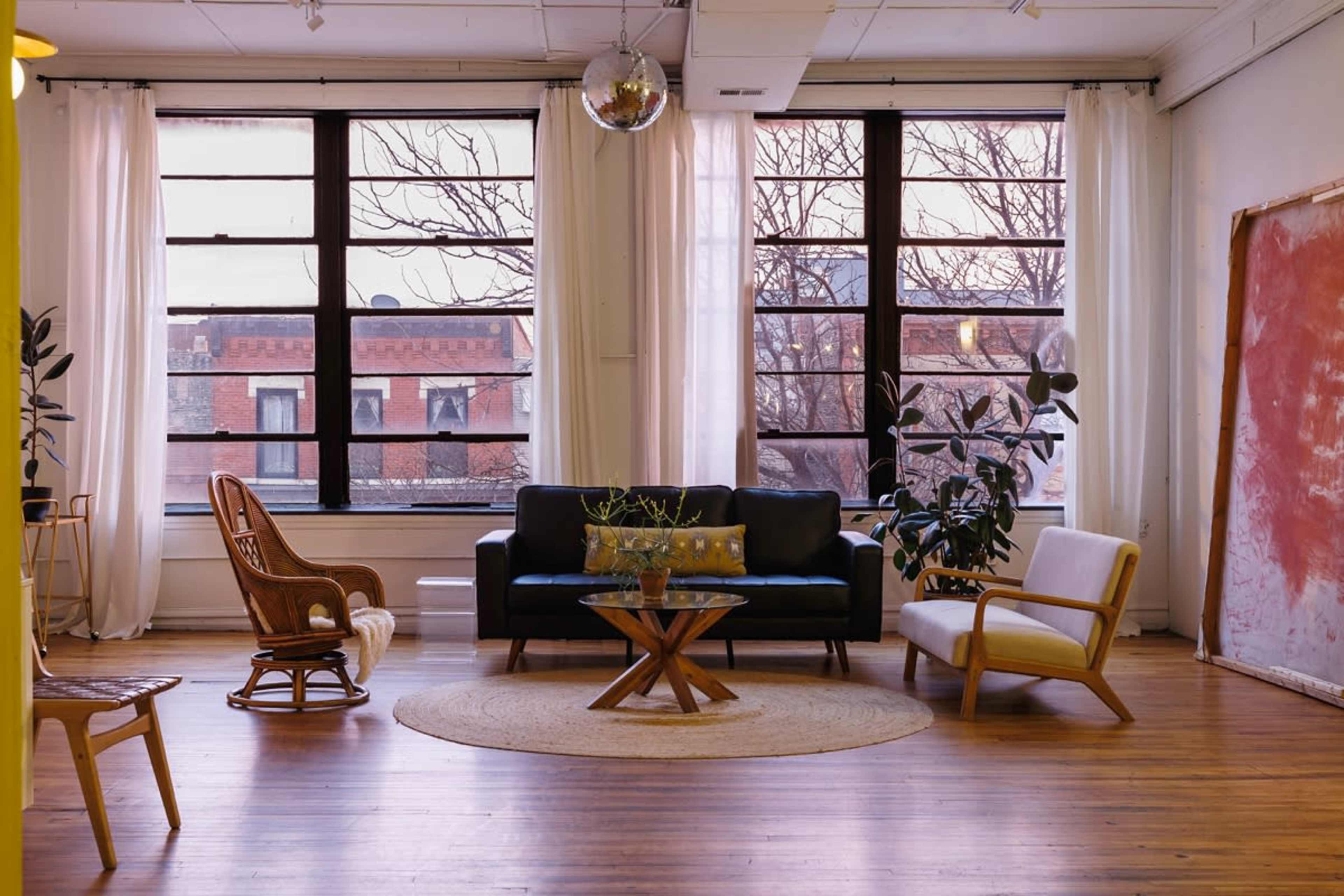A spacious living room features large windows, a black sofa, a white chair, a round wooden table, and a decorative rug on wooden flooring.
