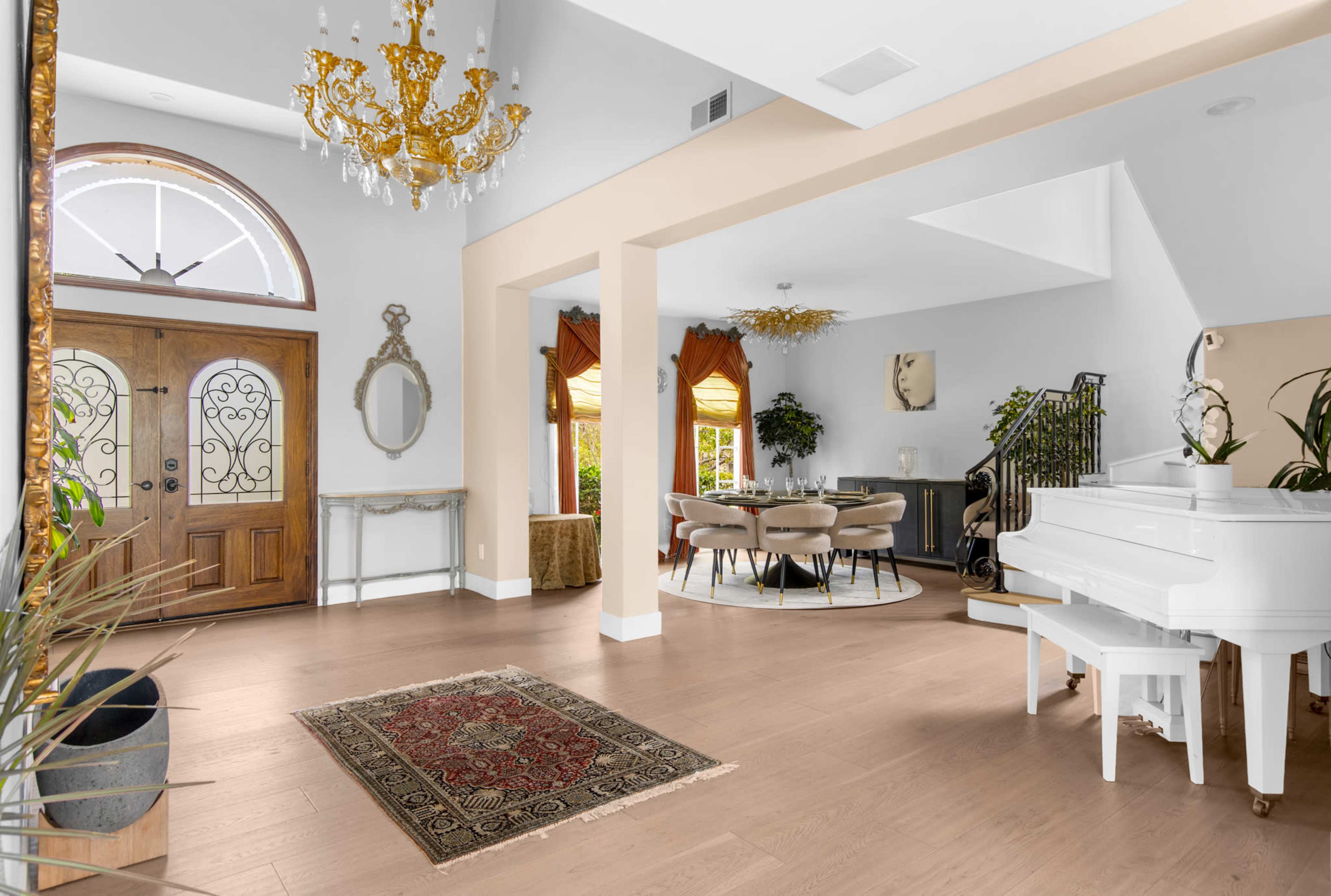The image shows a well-lit entryway featuring a grand chandelier, a white piano, and a circular dining table surrounded by chairs.