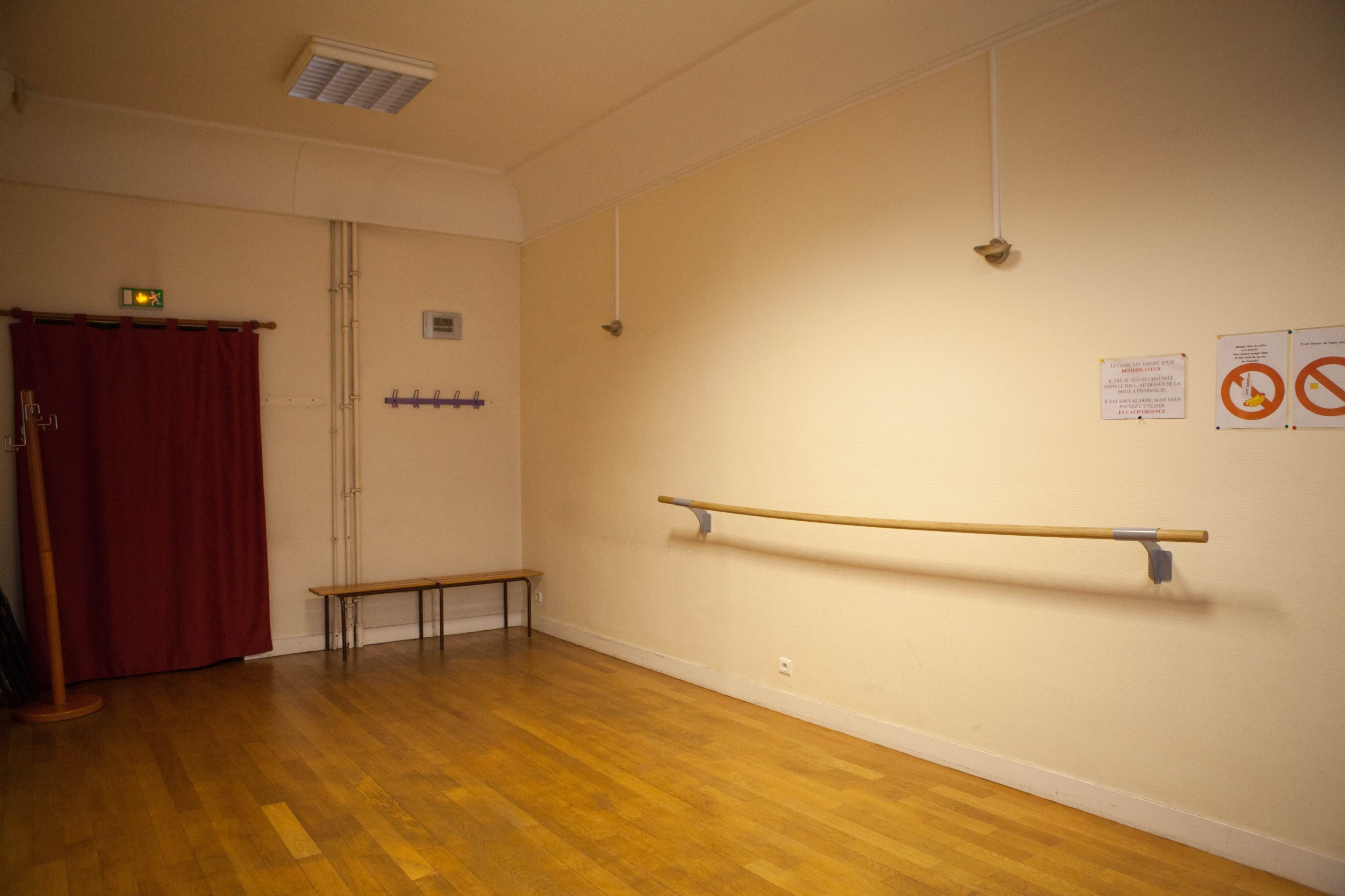 The image shows an empty dance studio with a wooden floor, a barre against the wall, a bench, and a curtain on one side.