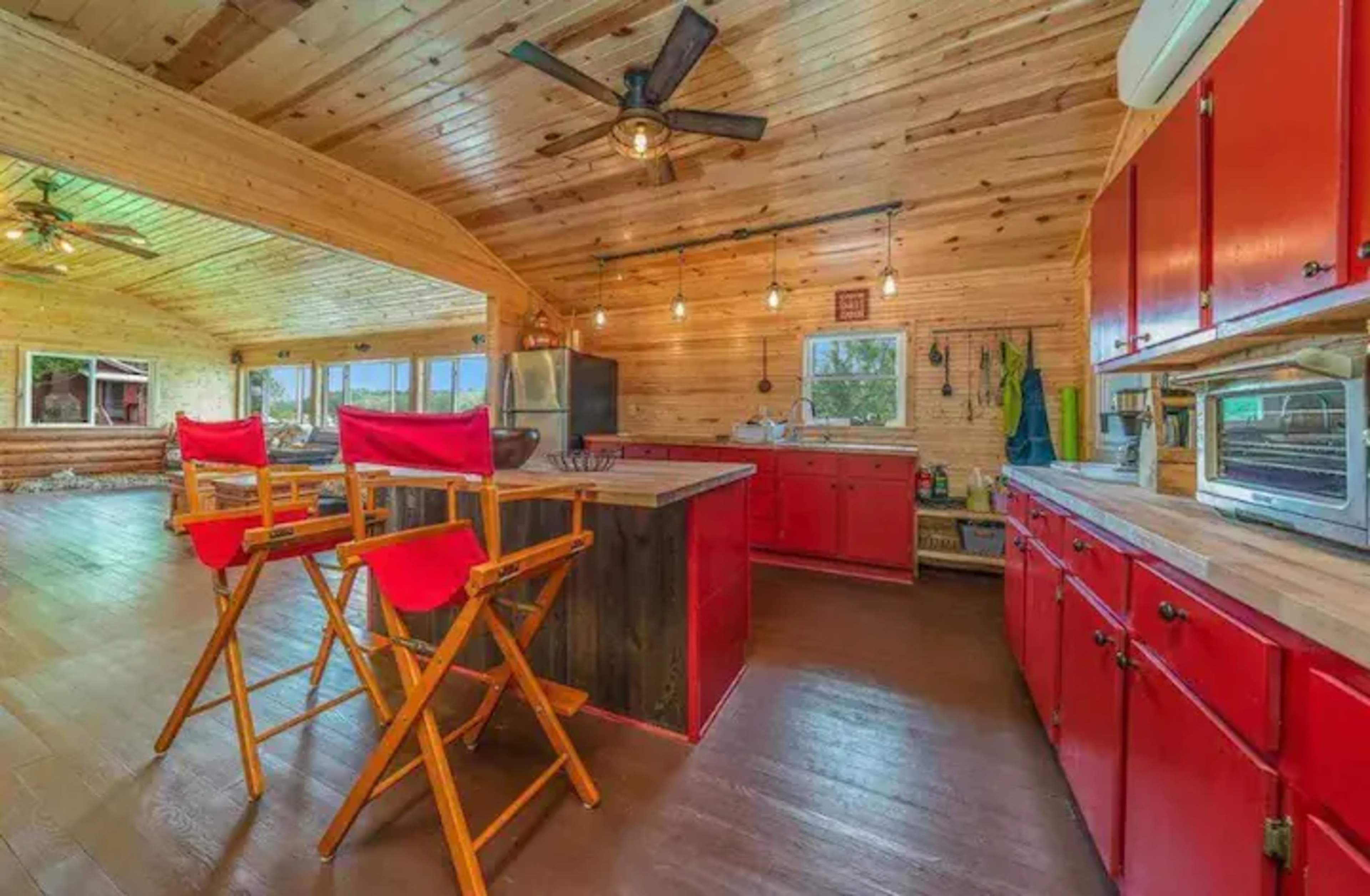 A rustic kitchen with wooden walls, red cabinets, and a large central island featuring two high-backed chairs.