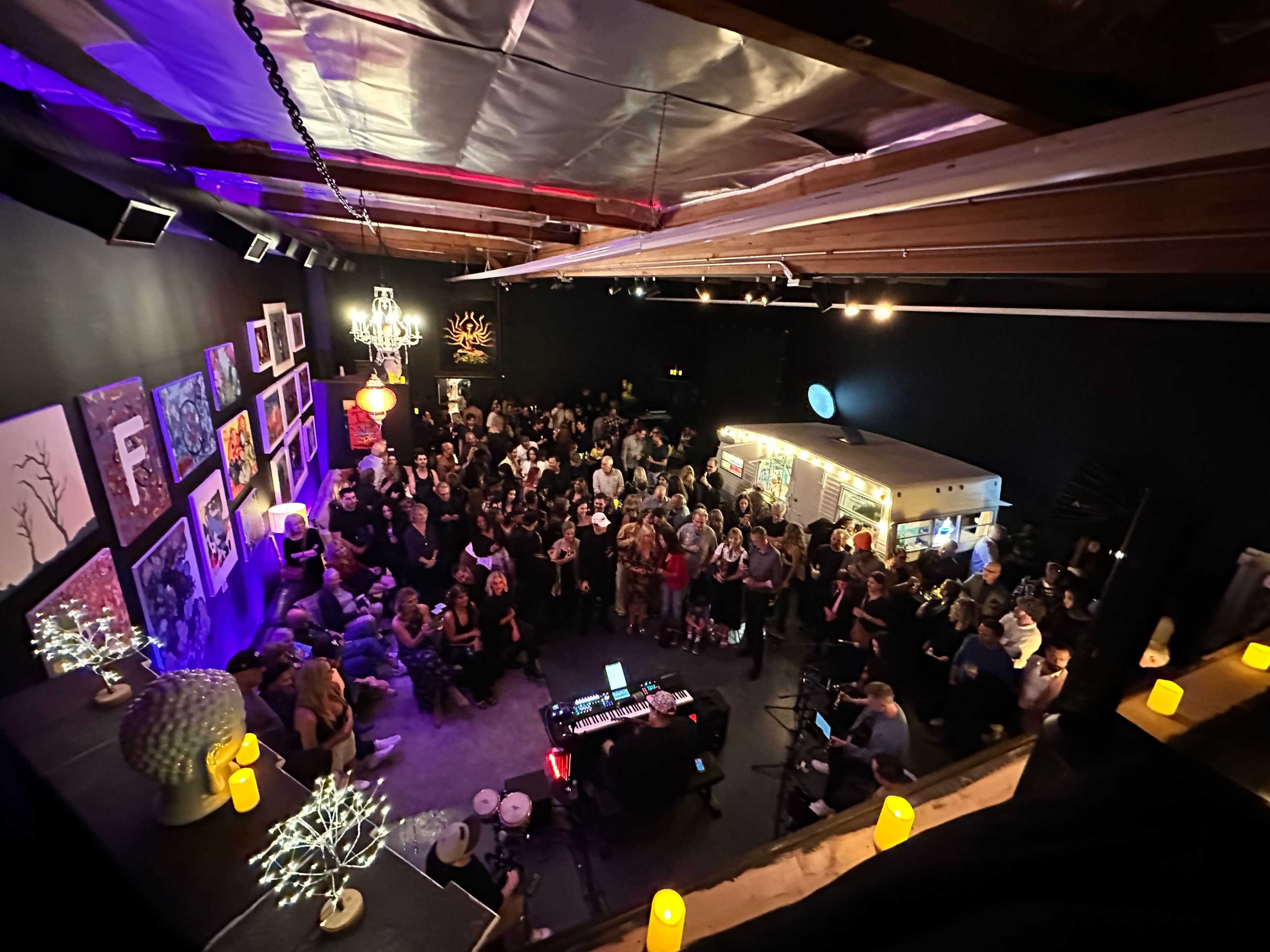 A large crowd gathers in a dimly lit venue filled with artwork, while a musician performs in front of them.