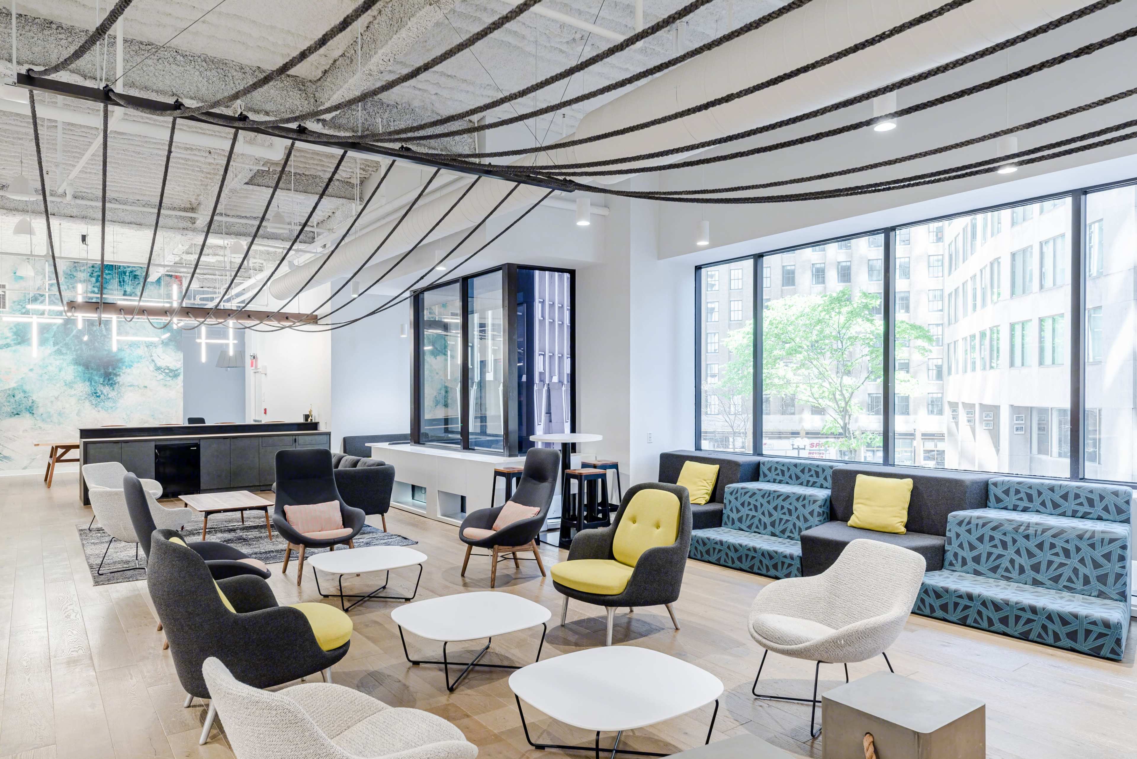 A modern office lounge area with various seating options, large windows, and suspended decorative elements above.