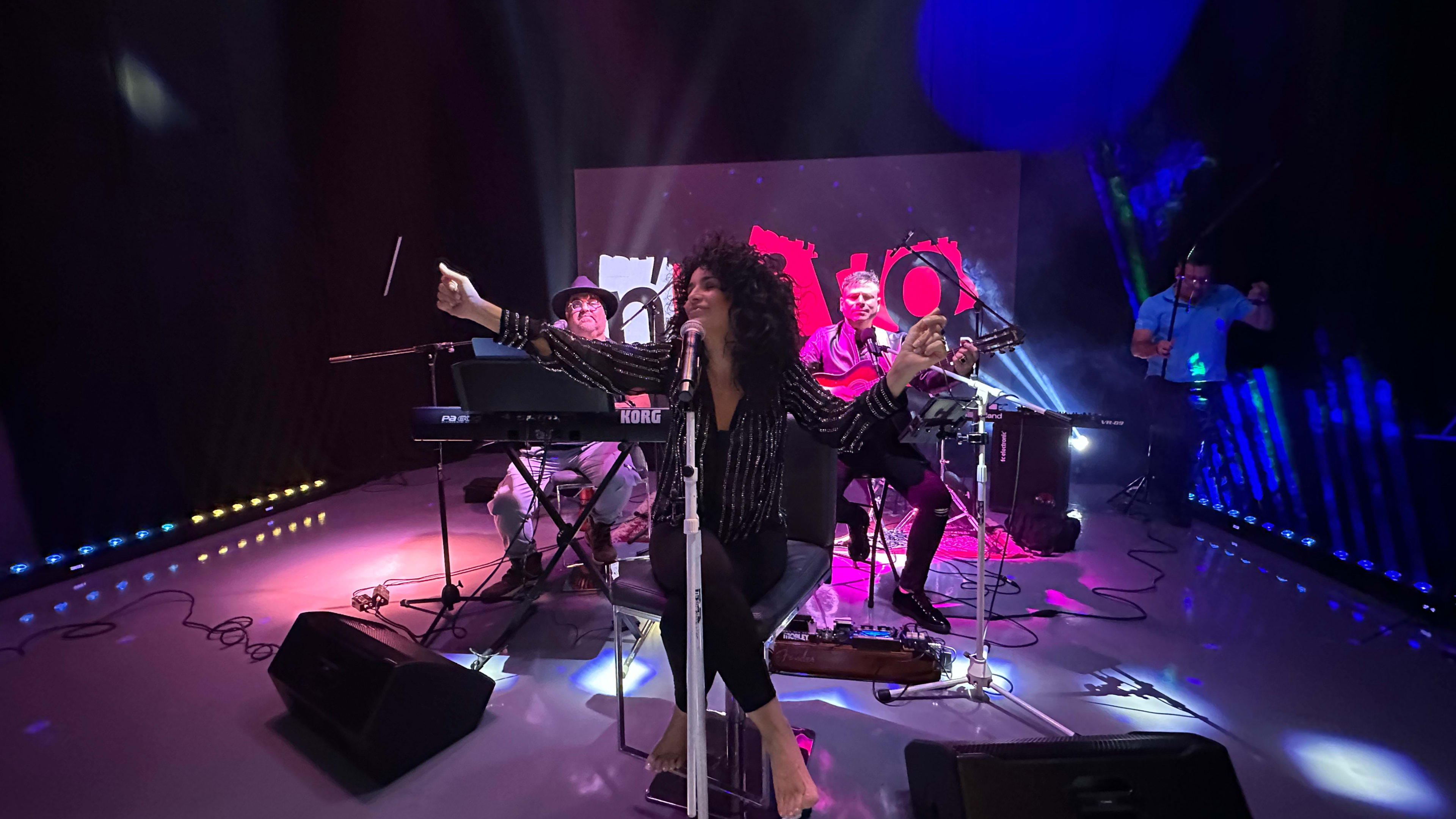 A musician performs at a studio with band members playing various instruments and colorful stage lighting in the background.