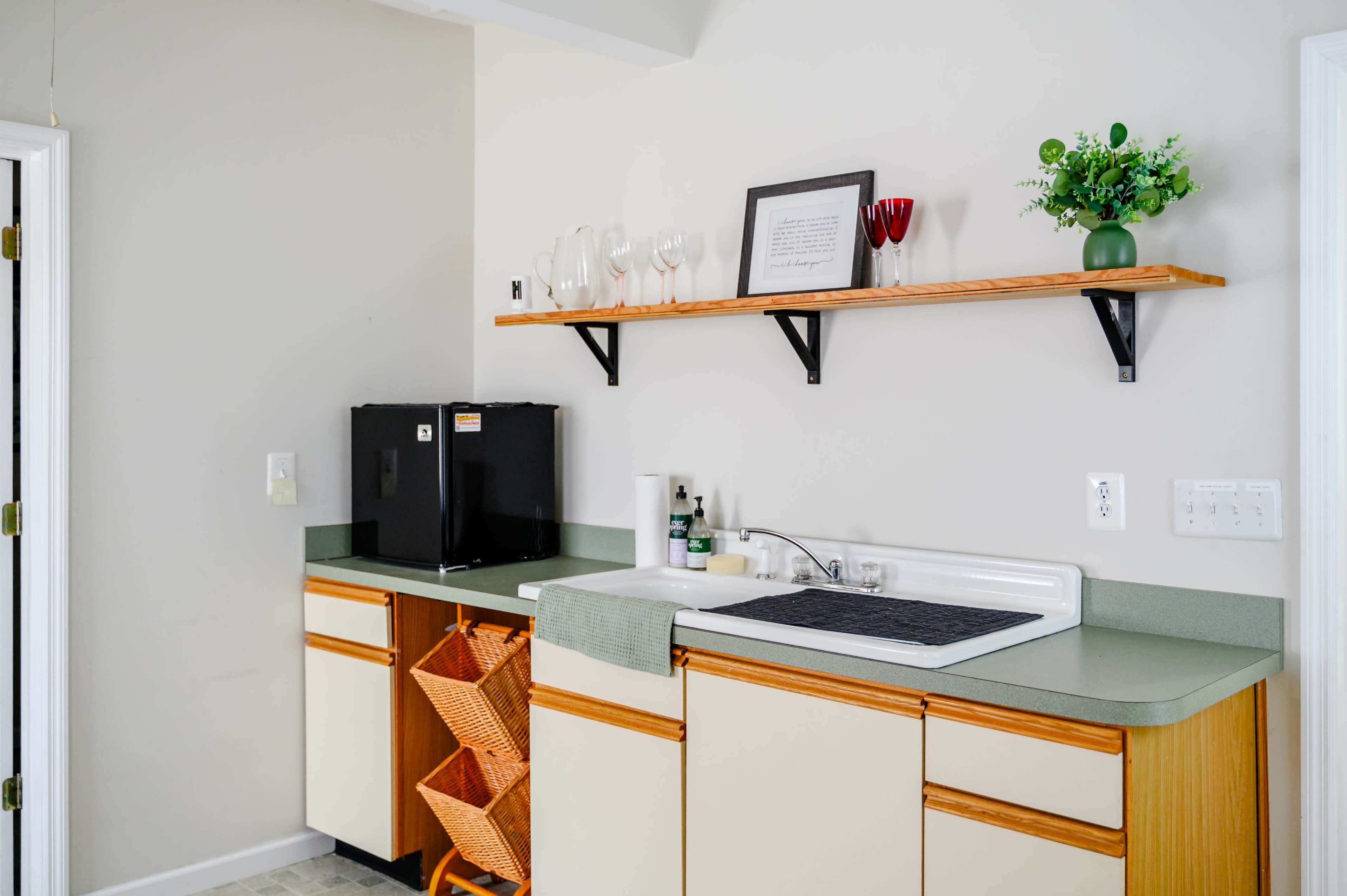 The image shows a small kitchen area featuring a sink, a countertop, and a shelf with glassware and a decorative plant, alongside a black mini-fridge and two wicker baskets for storage.