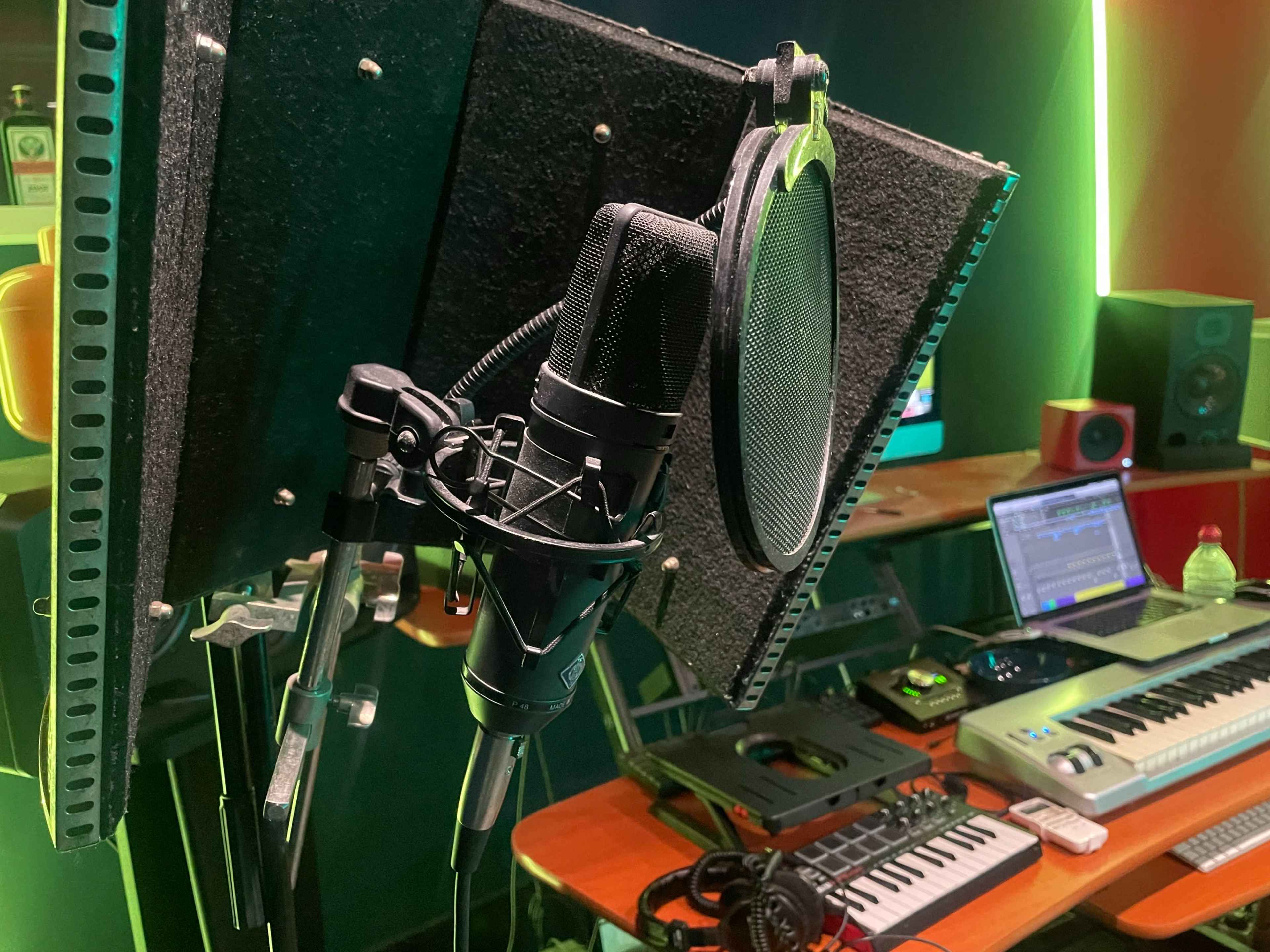 A microphone with a pop filter is positioned in a recording studio, surrounded by various music production equipment.