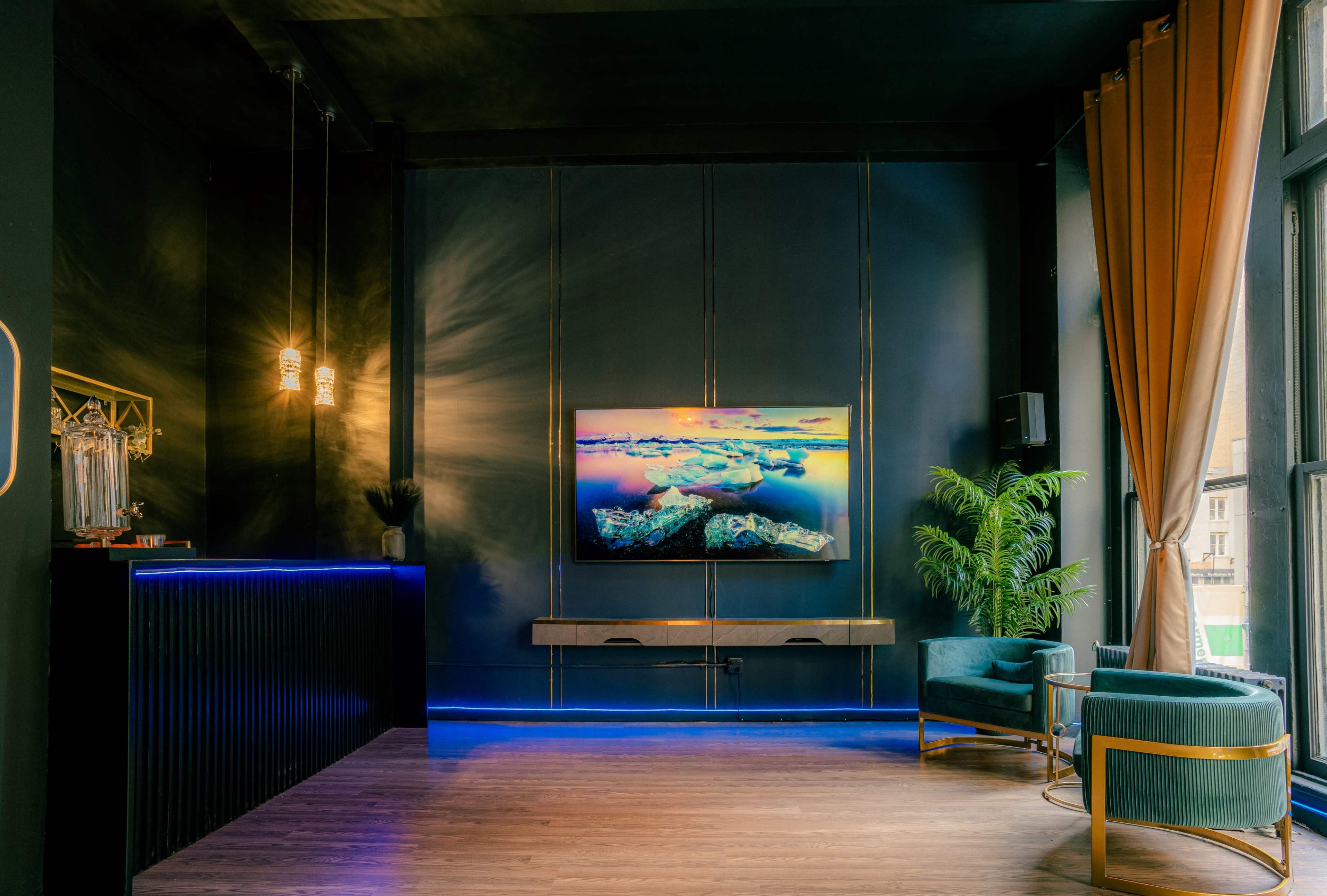 A modern reception area features a dark color scheme, a wall-mounted TV displaying a landscape, and stylish furniture including a green velvet chair and decorative plants.