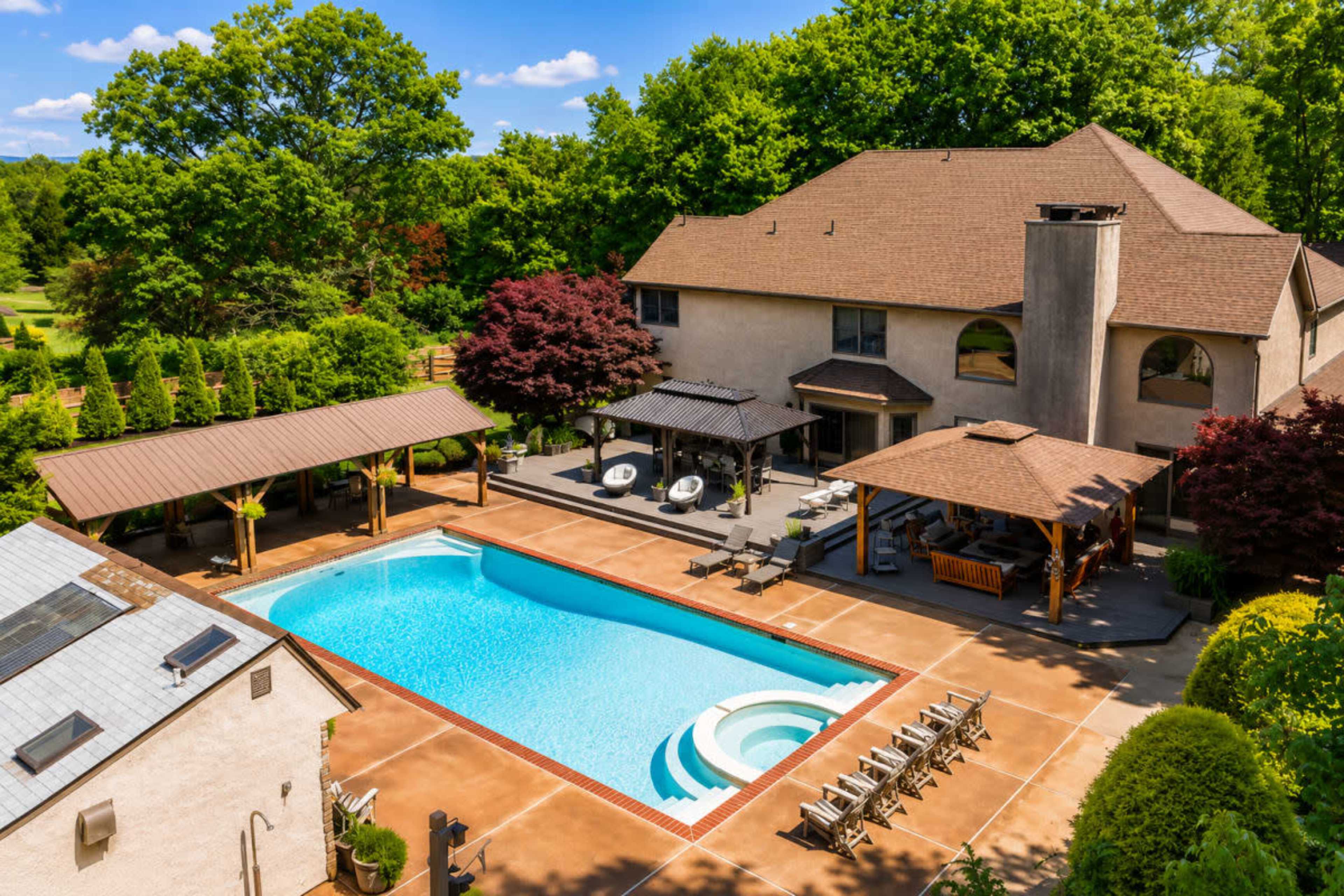 The image shows a spacious backyard featuring a large swimming pool, surrounded by lounge chairs and shaded areas, with a house and trees in the background.