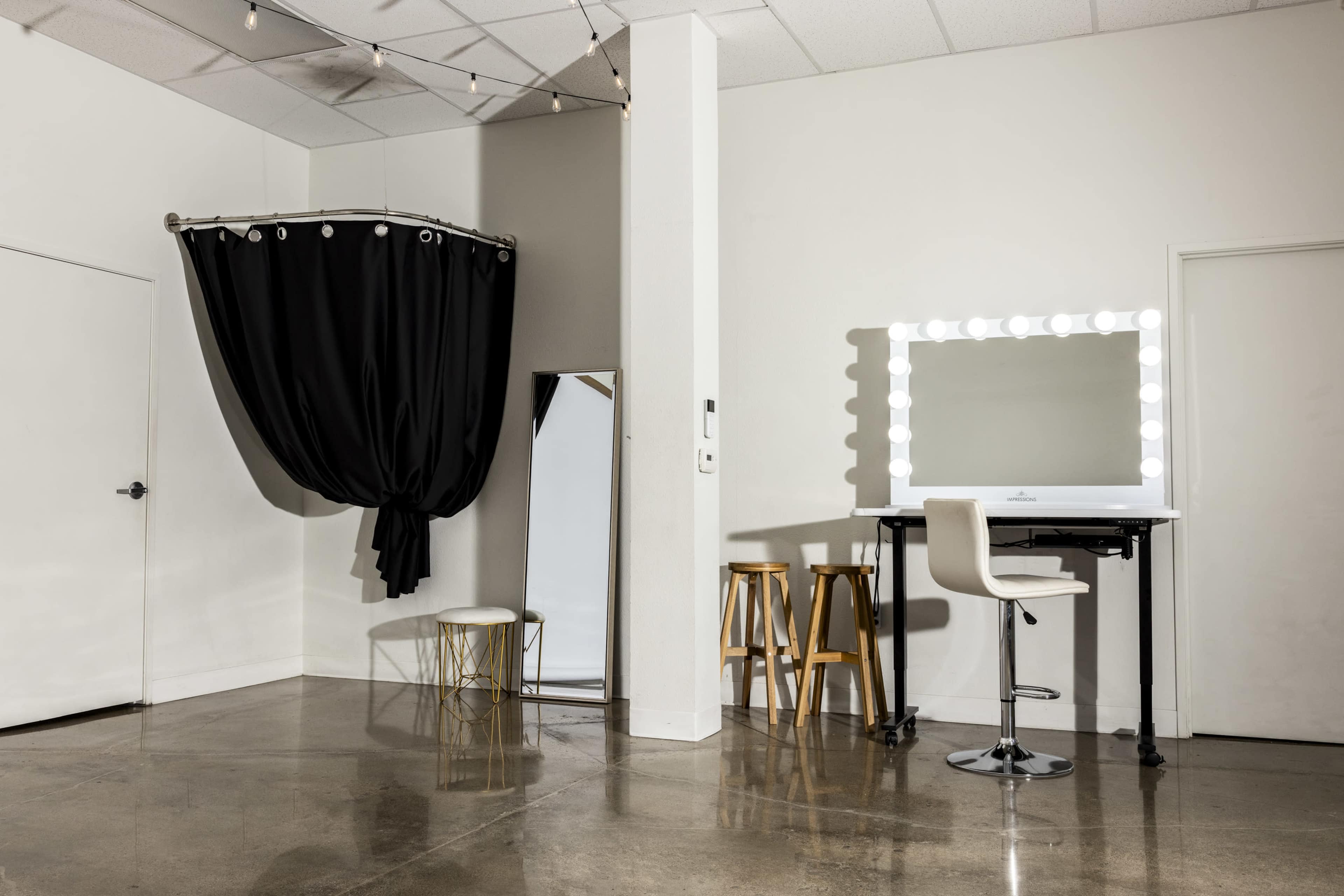 The image shows a minimalist room featuring a large mirror, a makeup station with bright lights, a stool, and a black curtain hanging from the ceiling.