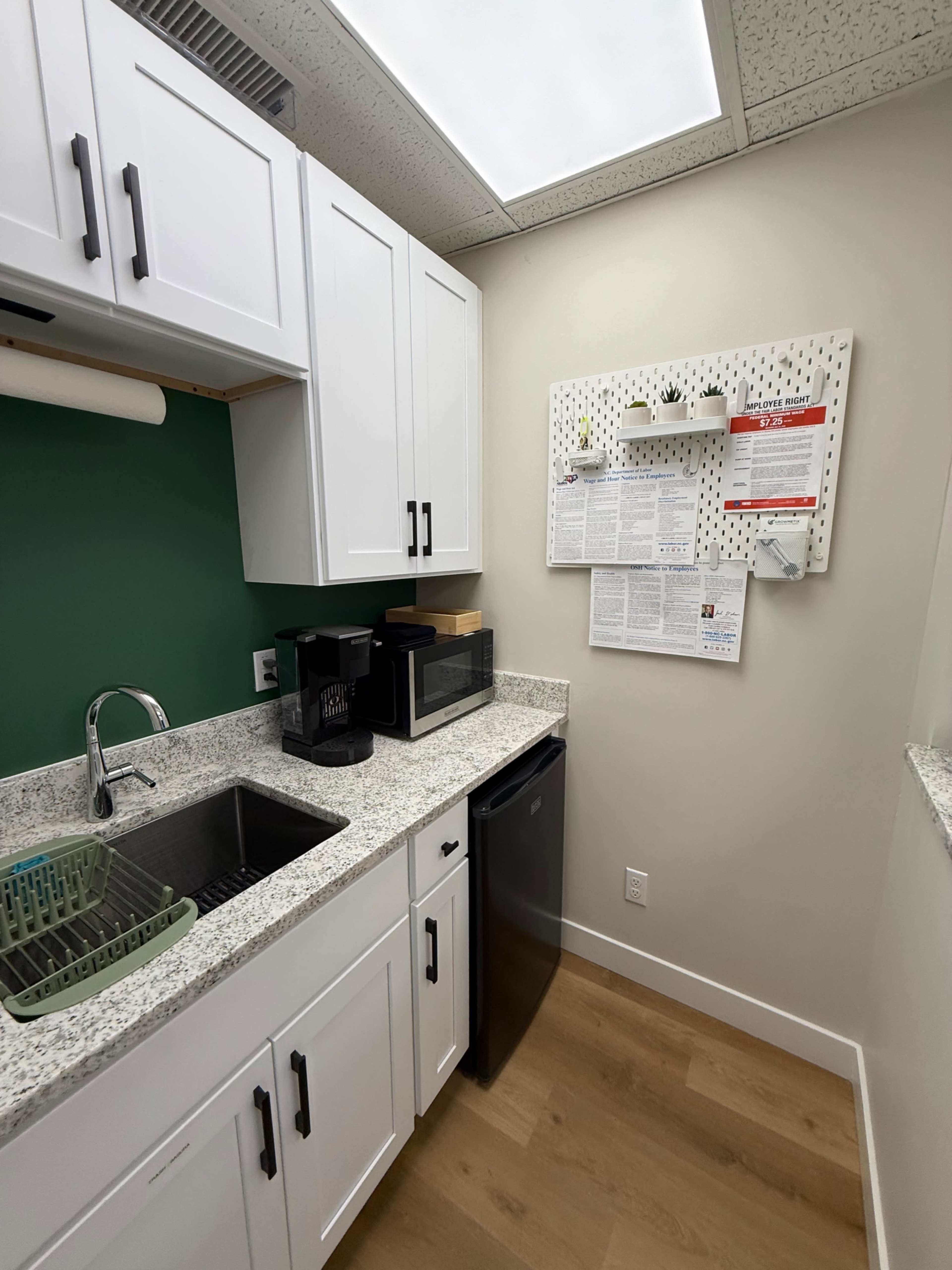 The image shows a small kitchen area featuring a sink, countertop, microwave, coffee maker, and a black refrigerator, along with a pegboard wall displaying various items and a calendar.