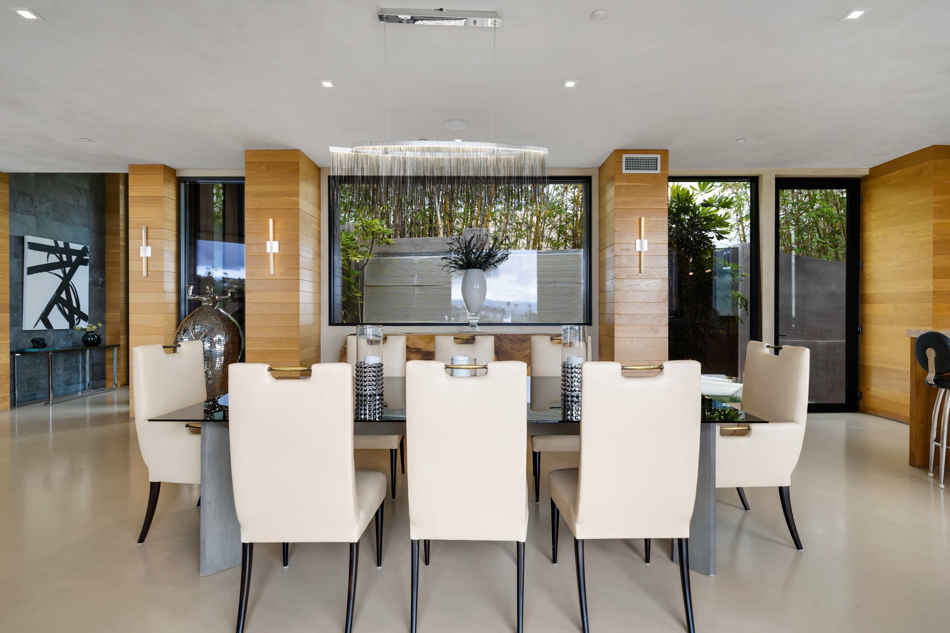 A dining room features a glass-topped table surrounded by cream-colored chairs, with large windows displaying greenery in the background.