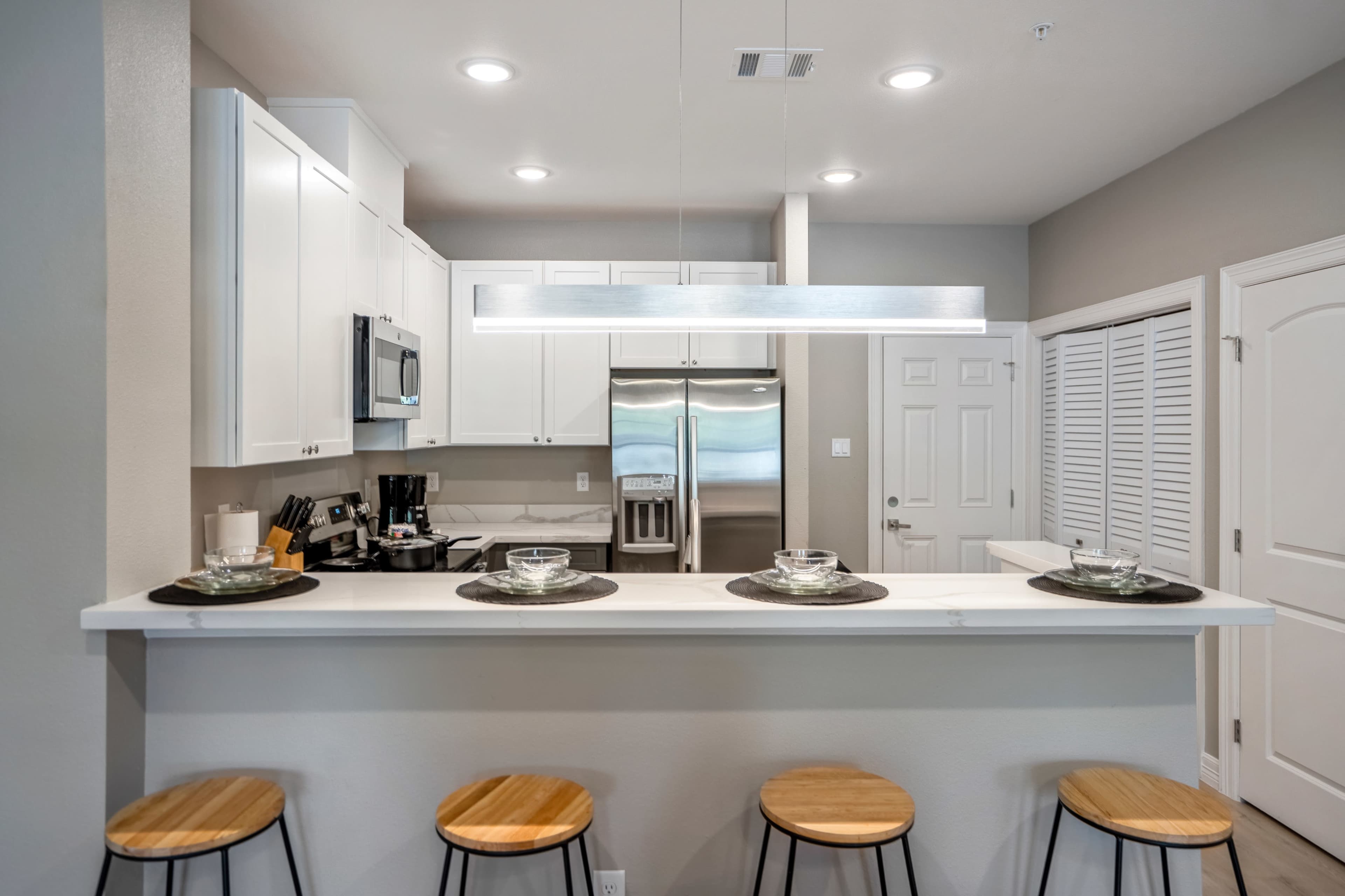 A modern kitchen features a counter with four wooden stools, sleek white cabinetry, and stainless steel appliances.