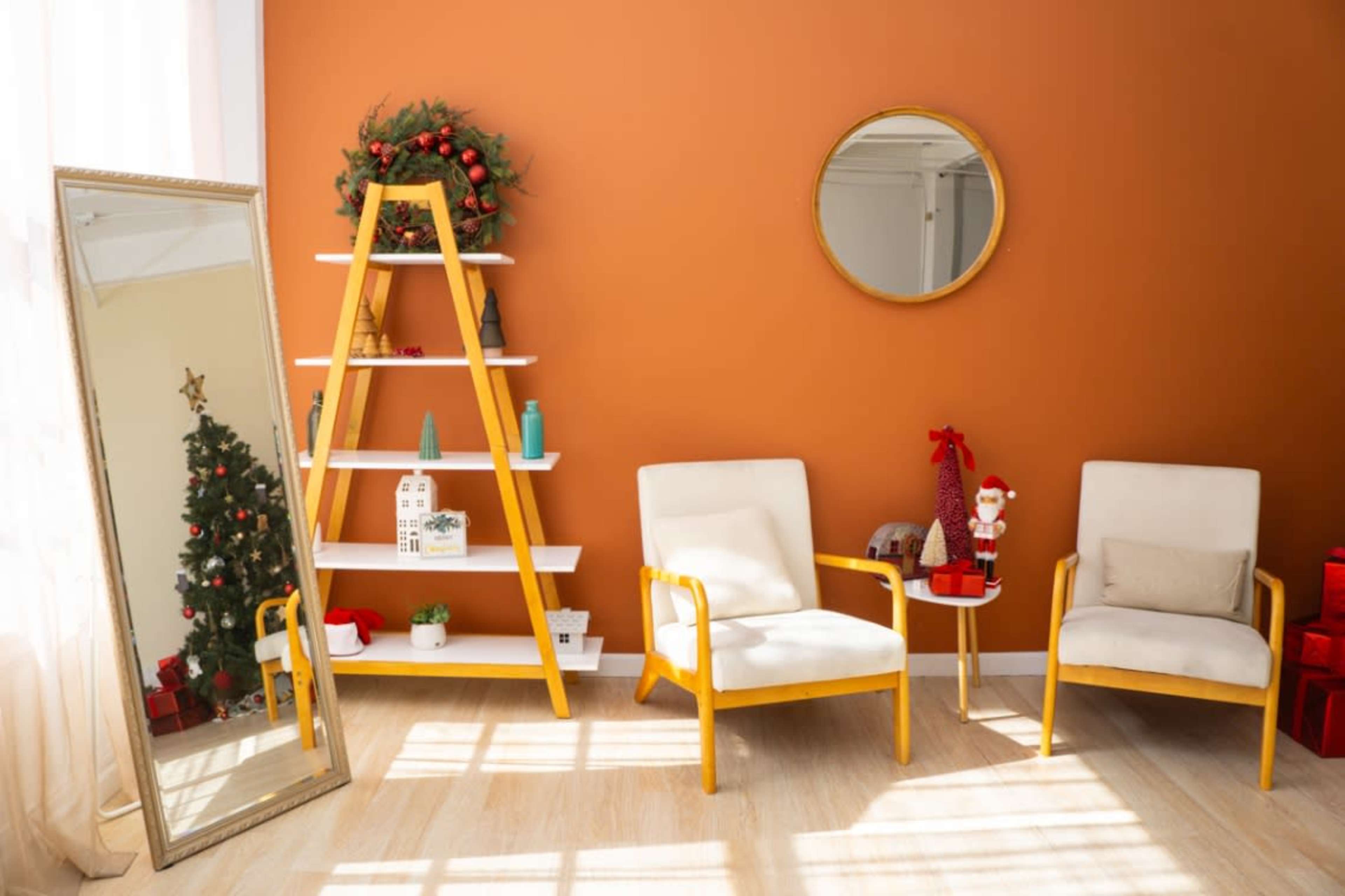 The image shows a cozy room with two light-colored chairs, a leaning mirror, a decorative ladder shelf, and a small Christmas tree near an orange wall.