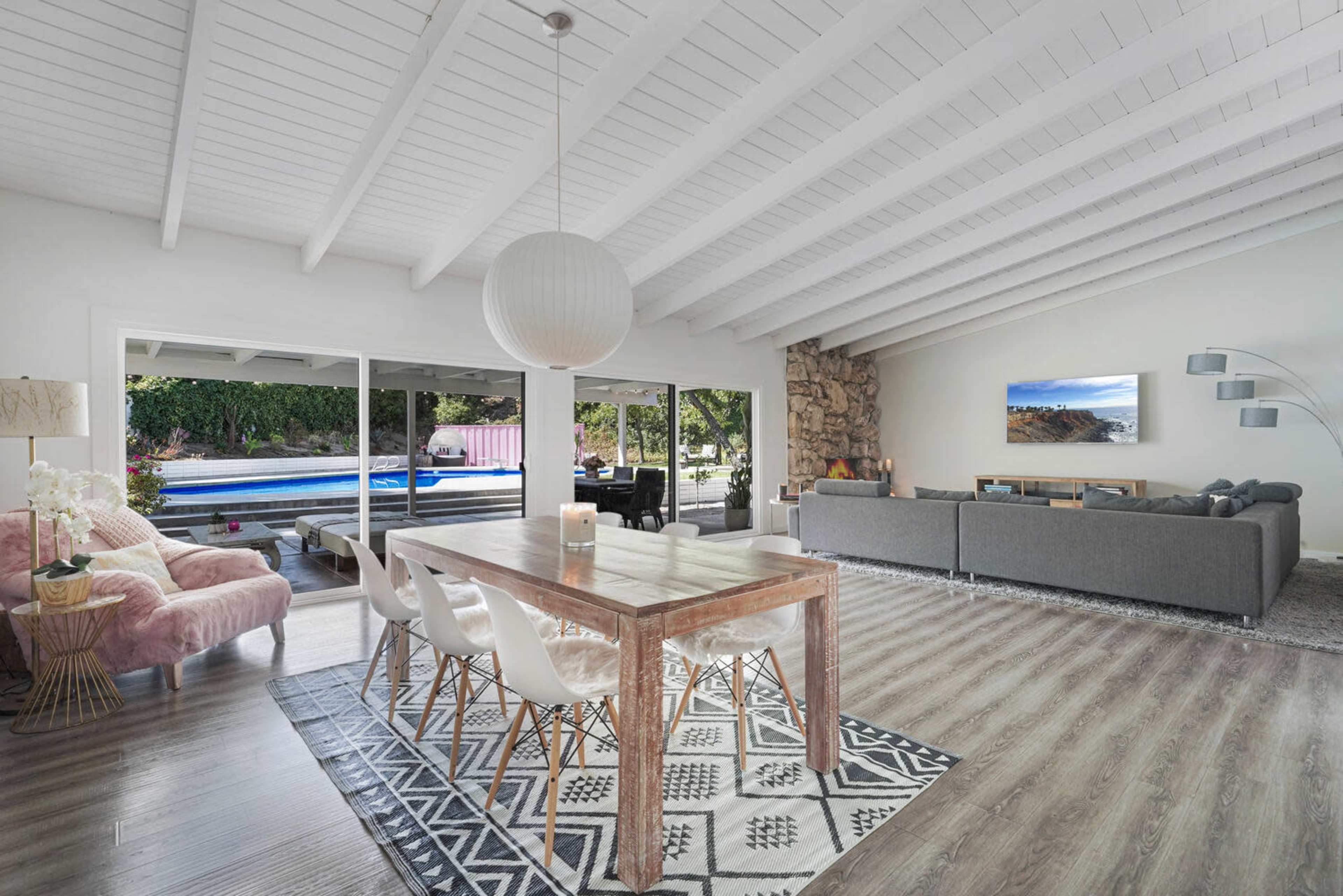 The scene shows a modern living room with a dining table, sofas, and large windows overlooking a pool area.