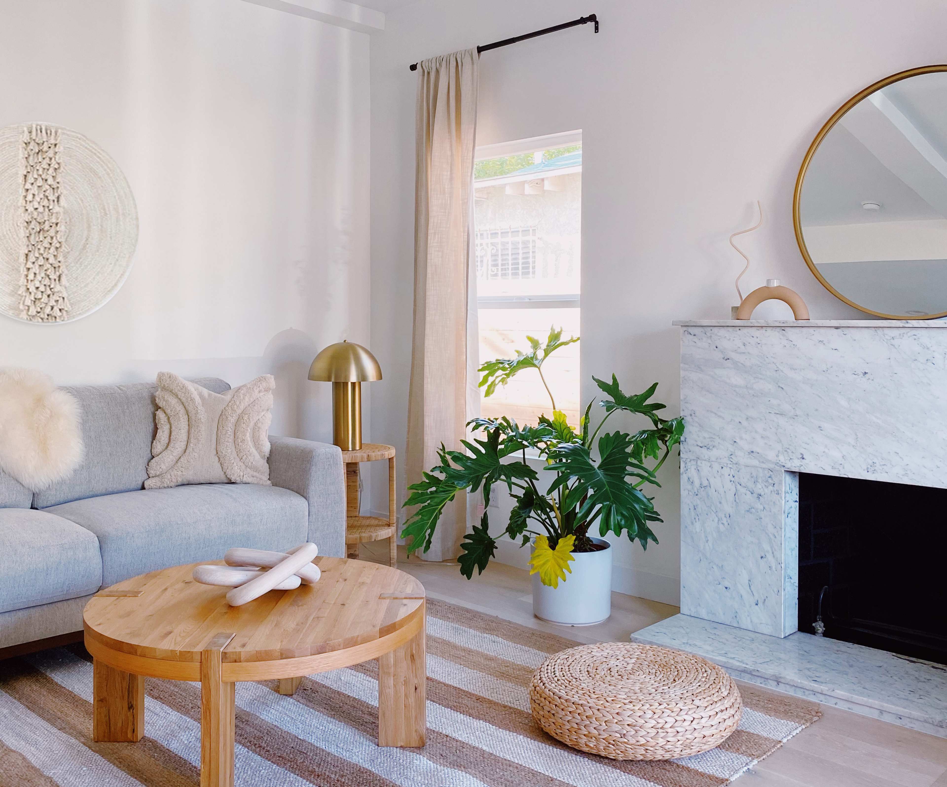 A light and airy living room features a gray sofa, a round wooden coffee table, a potted plant, and a decorative fireplace with a mirror above it.