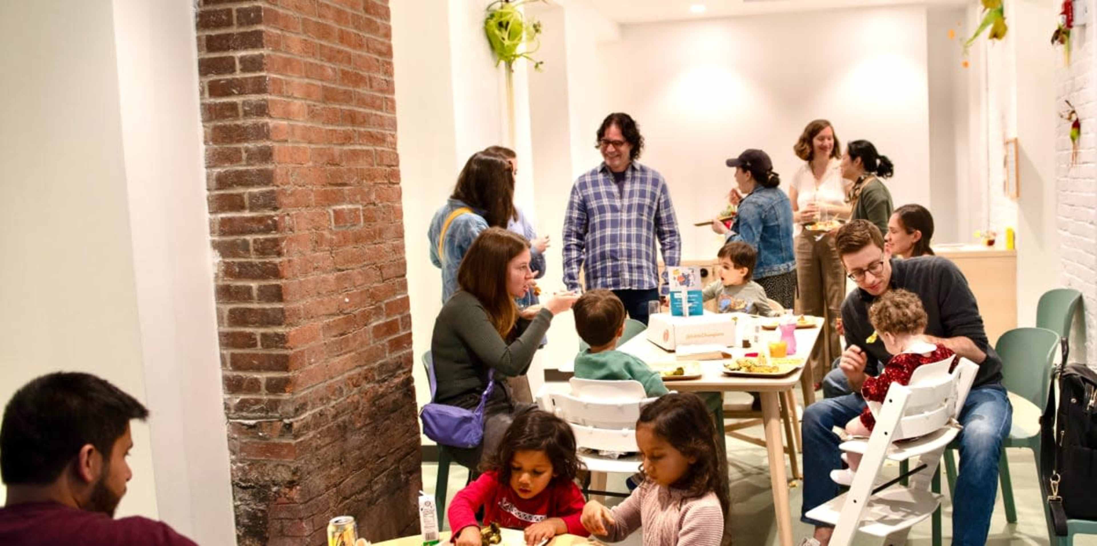 A group of adults and children are gathered around tables in a well-lit dining space, enjoying food and engaging in conversation.