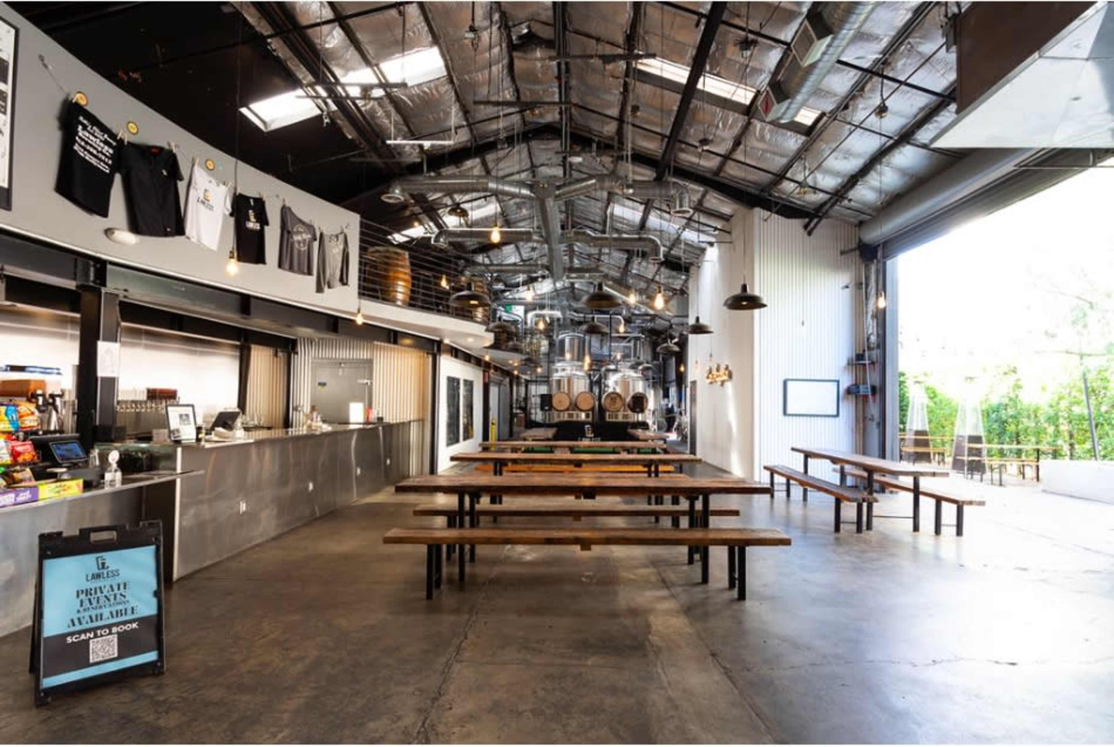 The image shows a spacious industrial-style dining area with long wooden tables, a counter with food service, and large windows that open to an outdoor area.