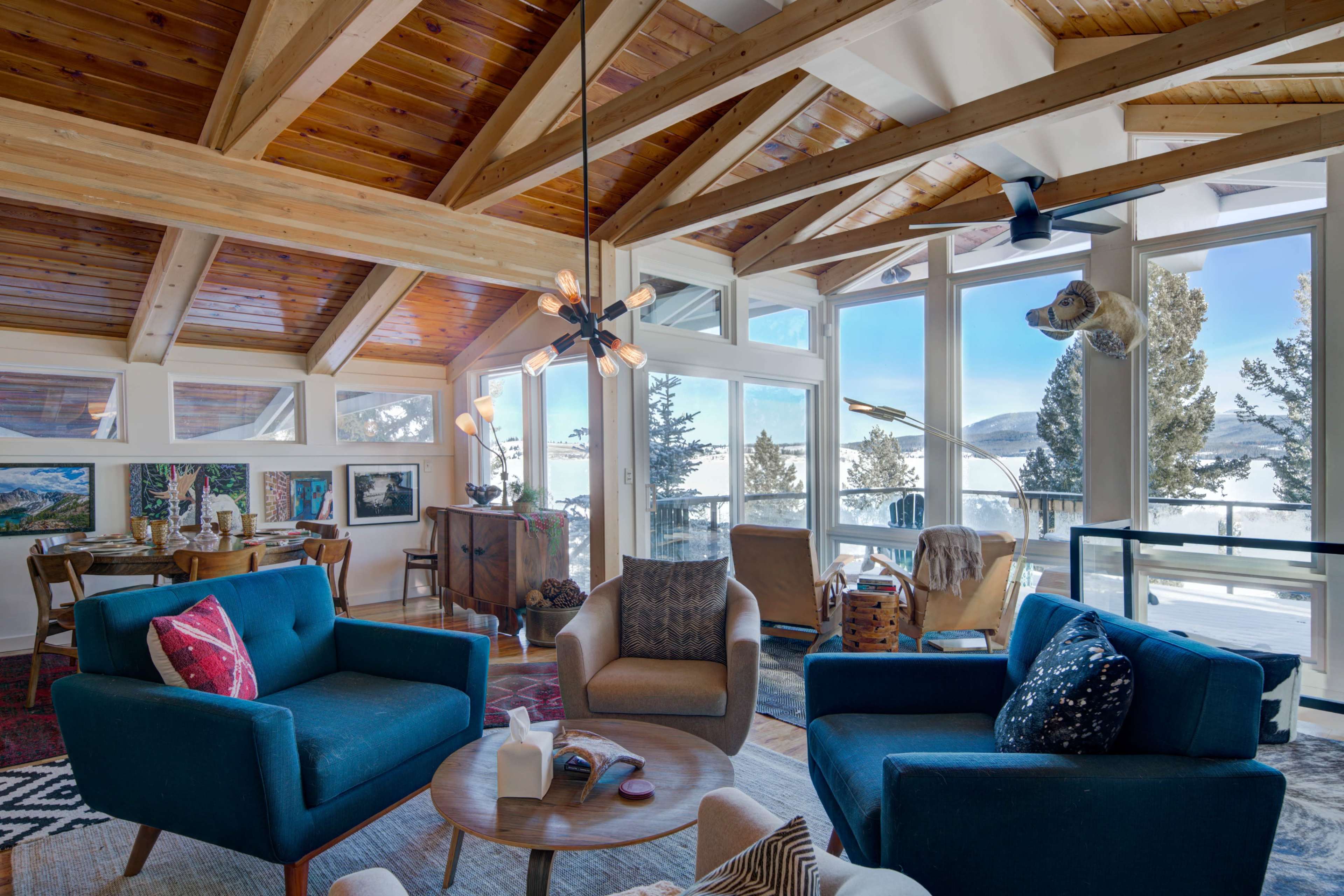 A spacious living room with modern furniture, large windows overlooking a snowy landscape, and exposed wooden beams on the ceiling.