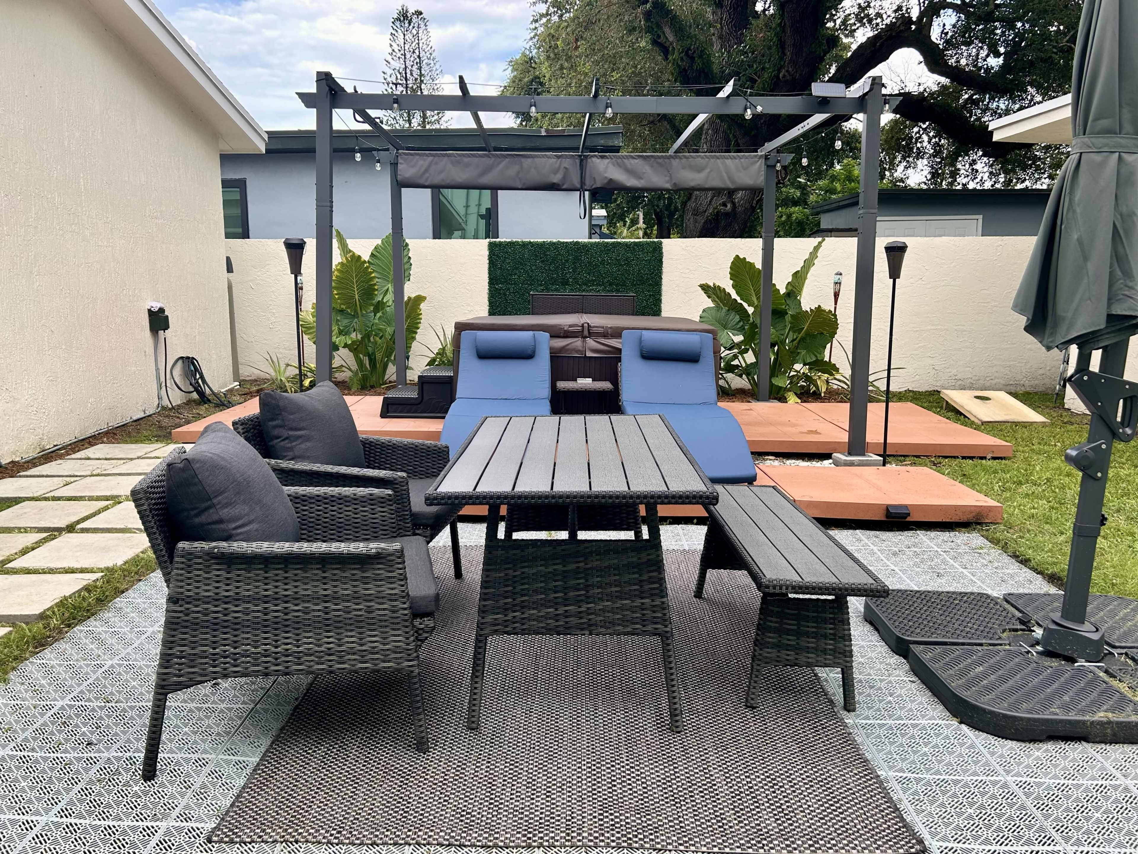 The image shows a patio area with a dining table and chairs, a couple of lounge chairs, and a covered seating space surrounded by greenery and paving stones.
