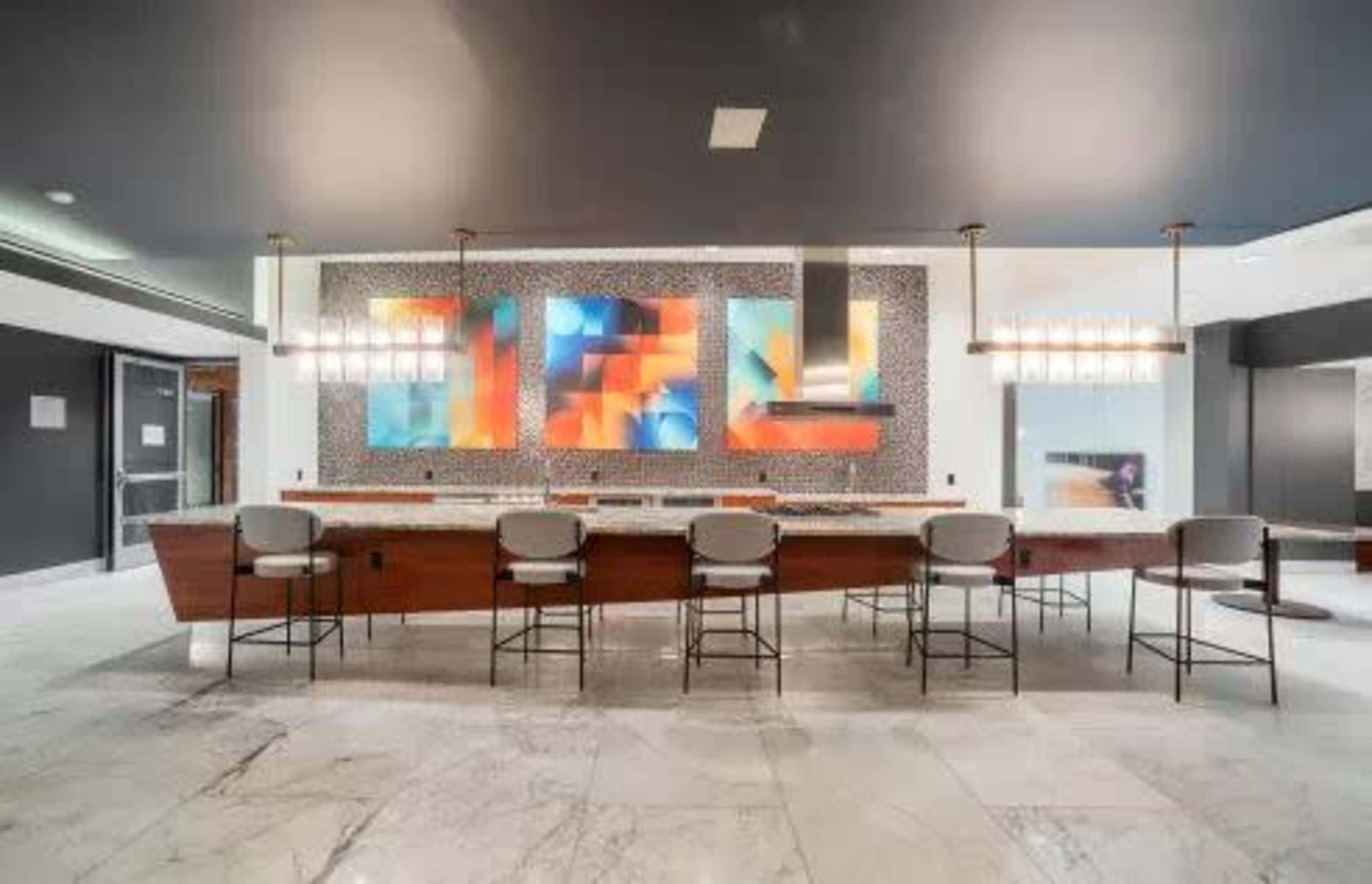 The image shows a modern bar area with a long wooden counter, four stools, and a colorful abstract painting on the wall behind it.